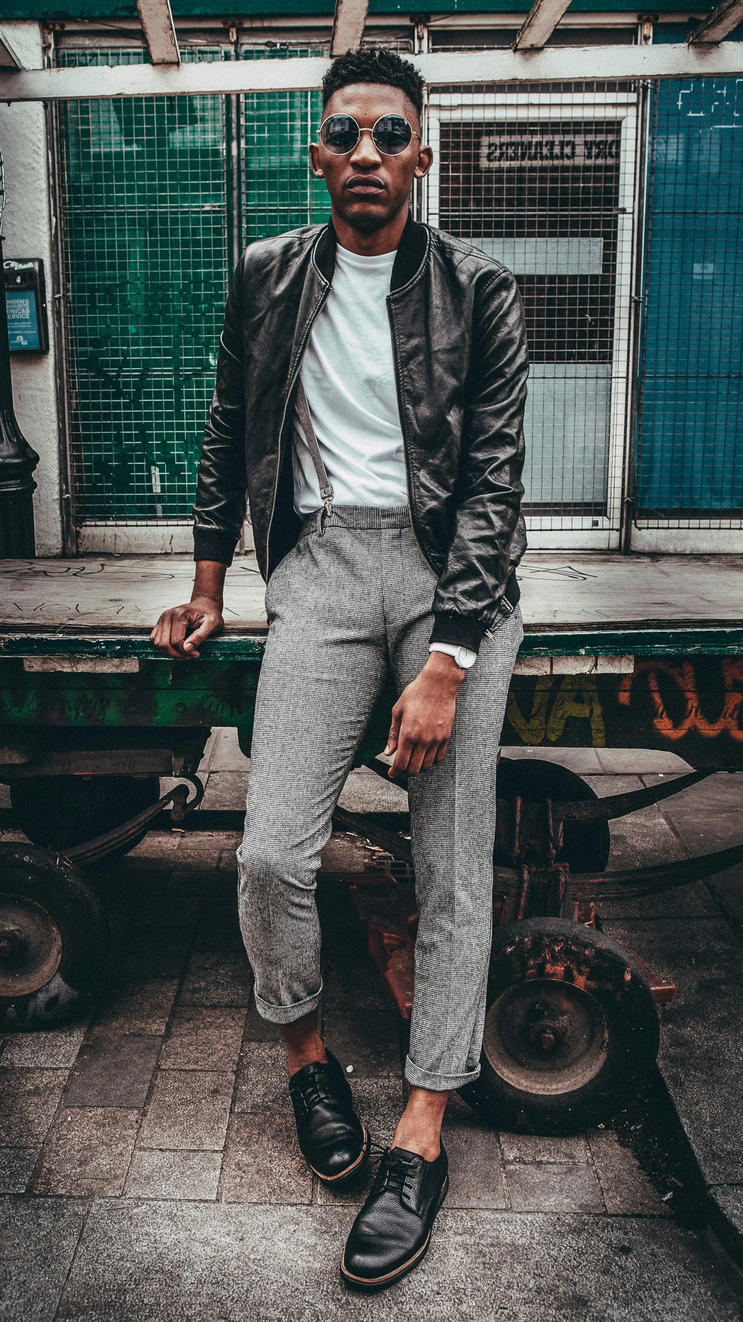 A young man with dark skin and short curly hair poses in front of a metal fence, wearing round sunglasses, a black leather jacket, a white T-shirt, gray patterned pants, black shoes, and a white watch on his left wrist. He stands on a paved sidewalk, with one hand resting on a green, graffiti-covered cart.