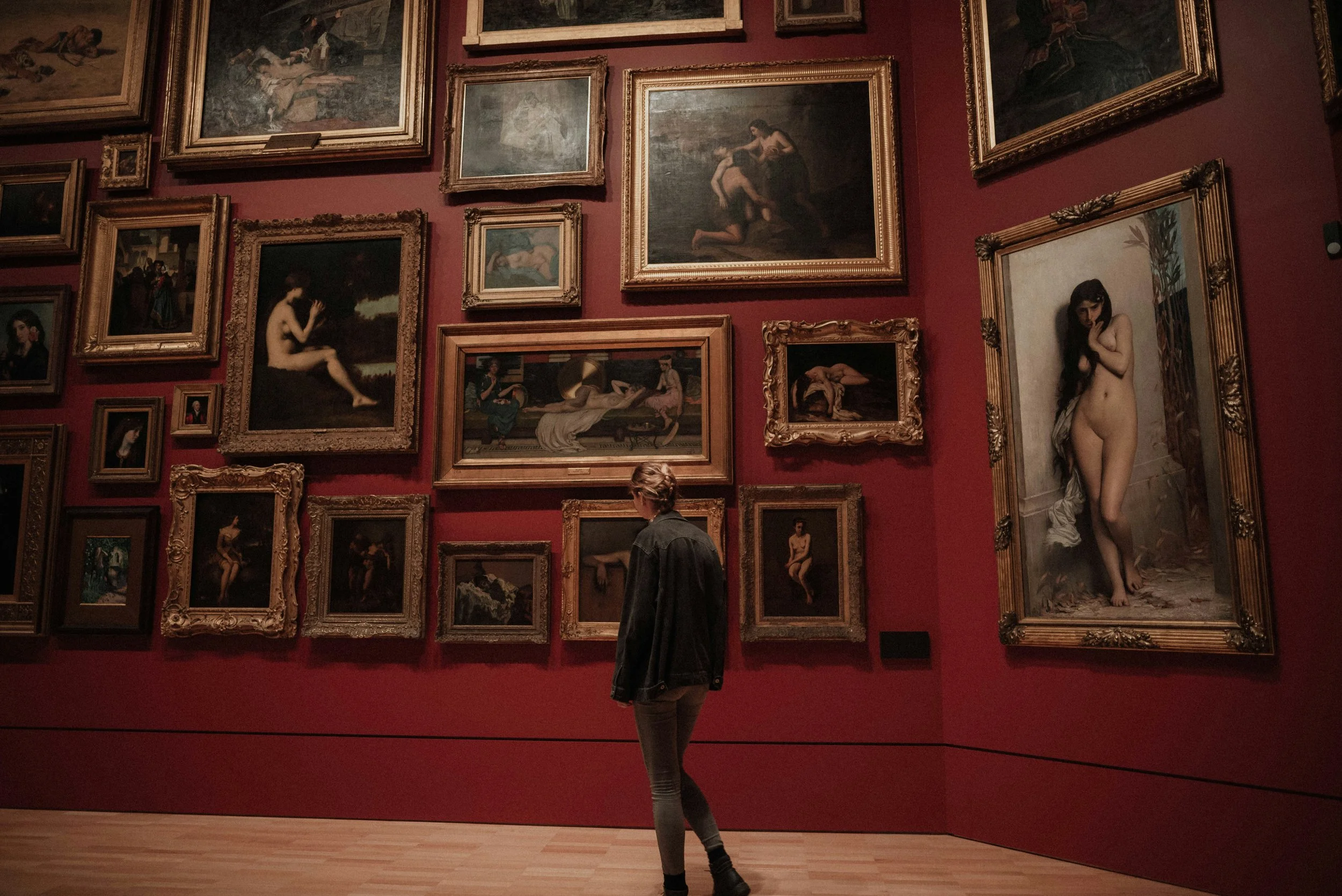 A person looking at framed paintings in an art gallery with a red wall and wooden floor.