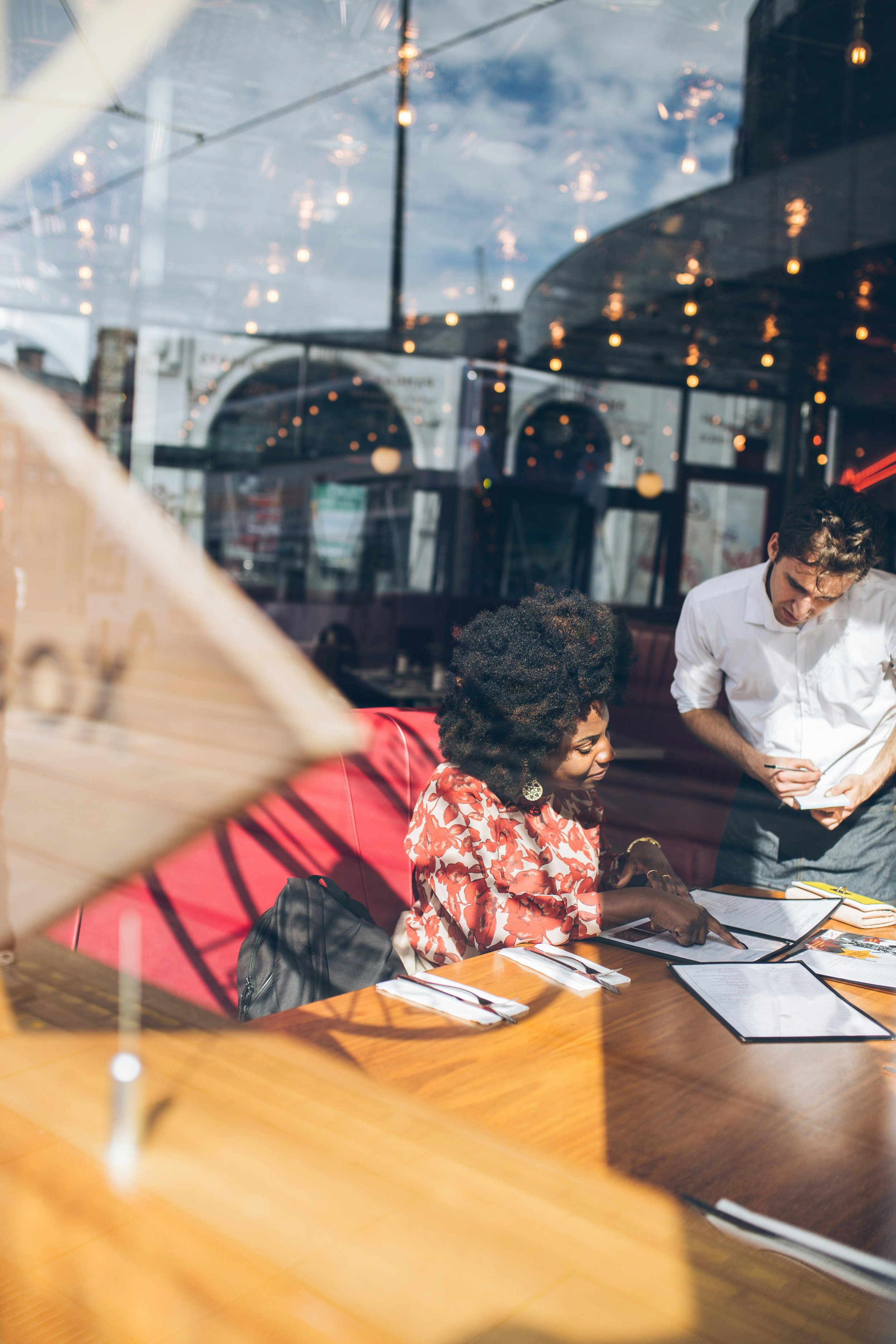 People sitting at a wooden table inside a restaurant, with reflections of string lights and an outdoor scene visible in the window behind them.