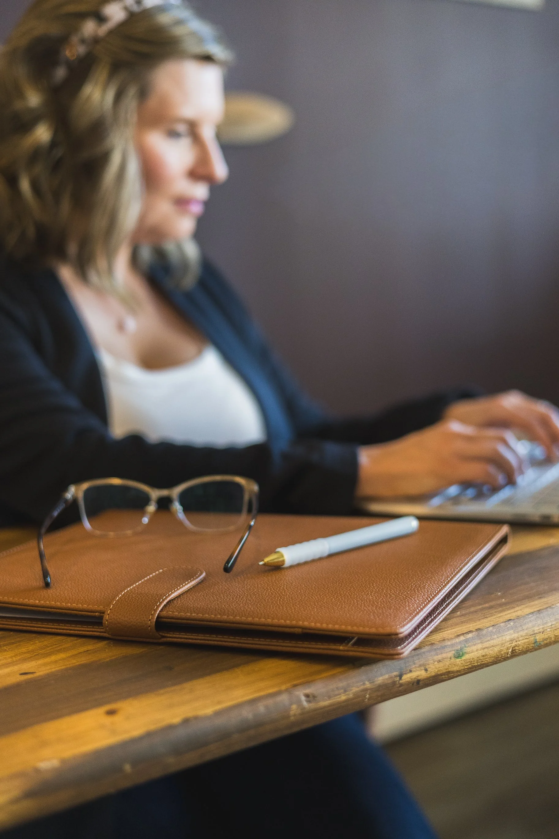A woman with wavy blonde hair sitting at a wooden table, working on a laptop. On the table, there are a pair of glasses, a white pen with a gold tip, and a brown leather folder.