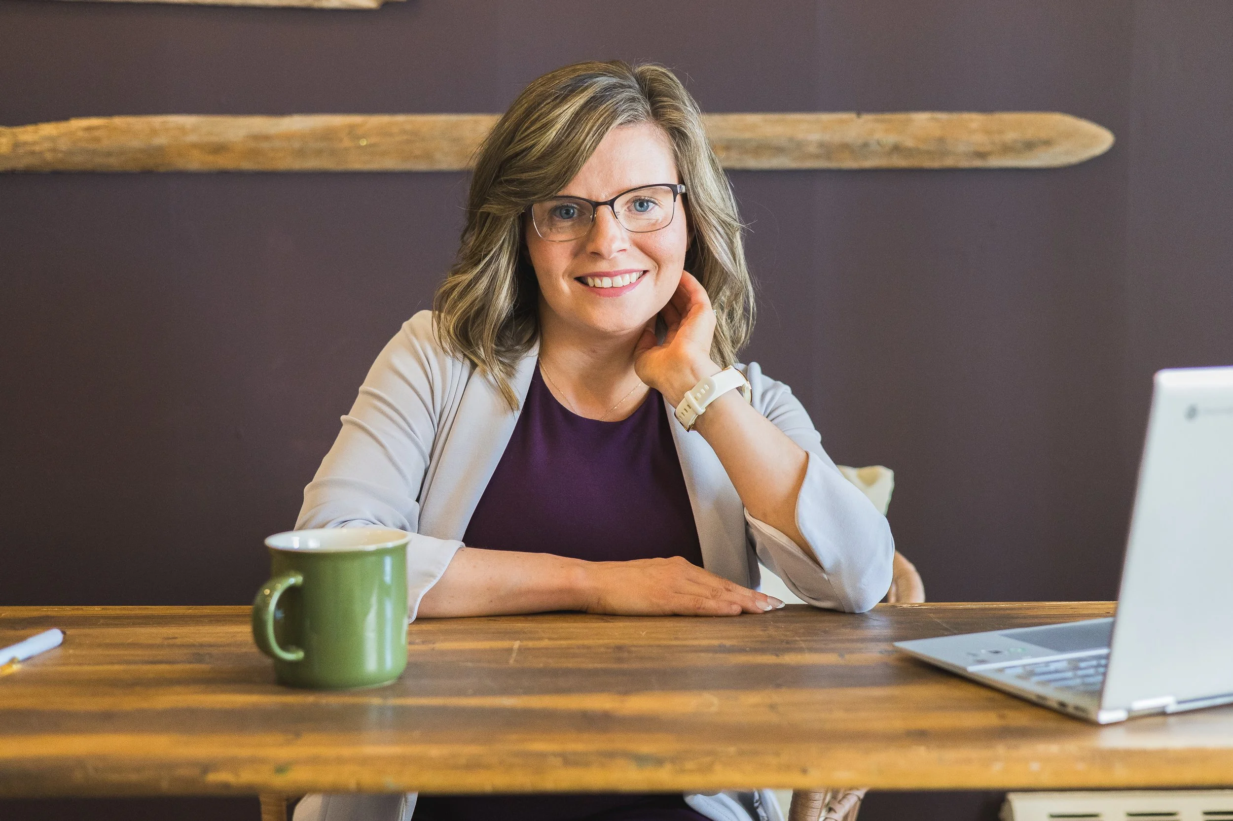 A woman with blonde hair and glasses sitting at a wooden table with a green mug and a laptop in front of her, smiling at the camera.