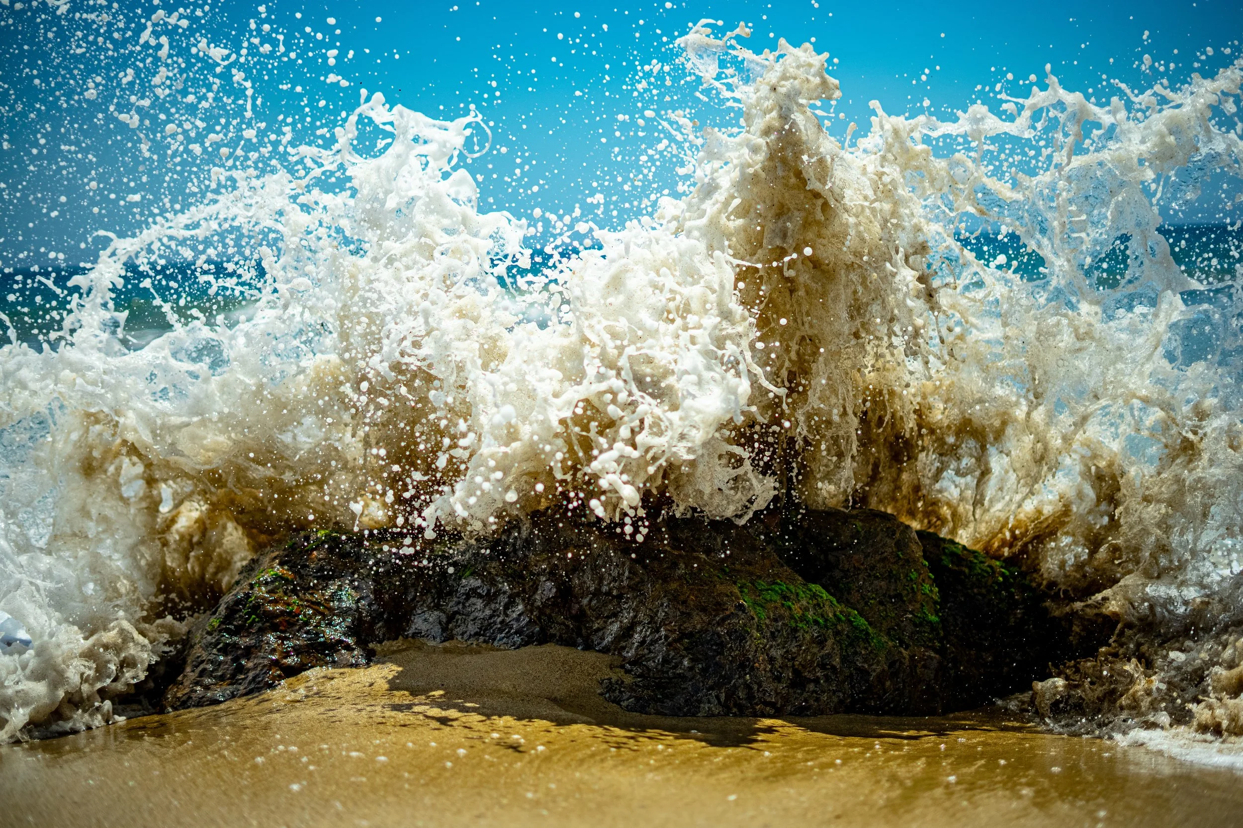 Waves crashing against rocks on a sandy beach under a clear blue sky.