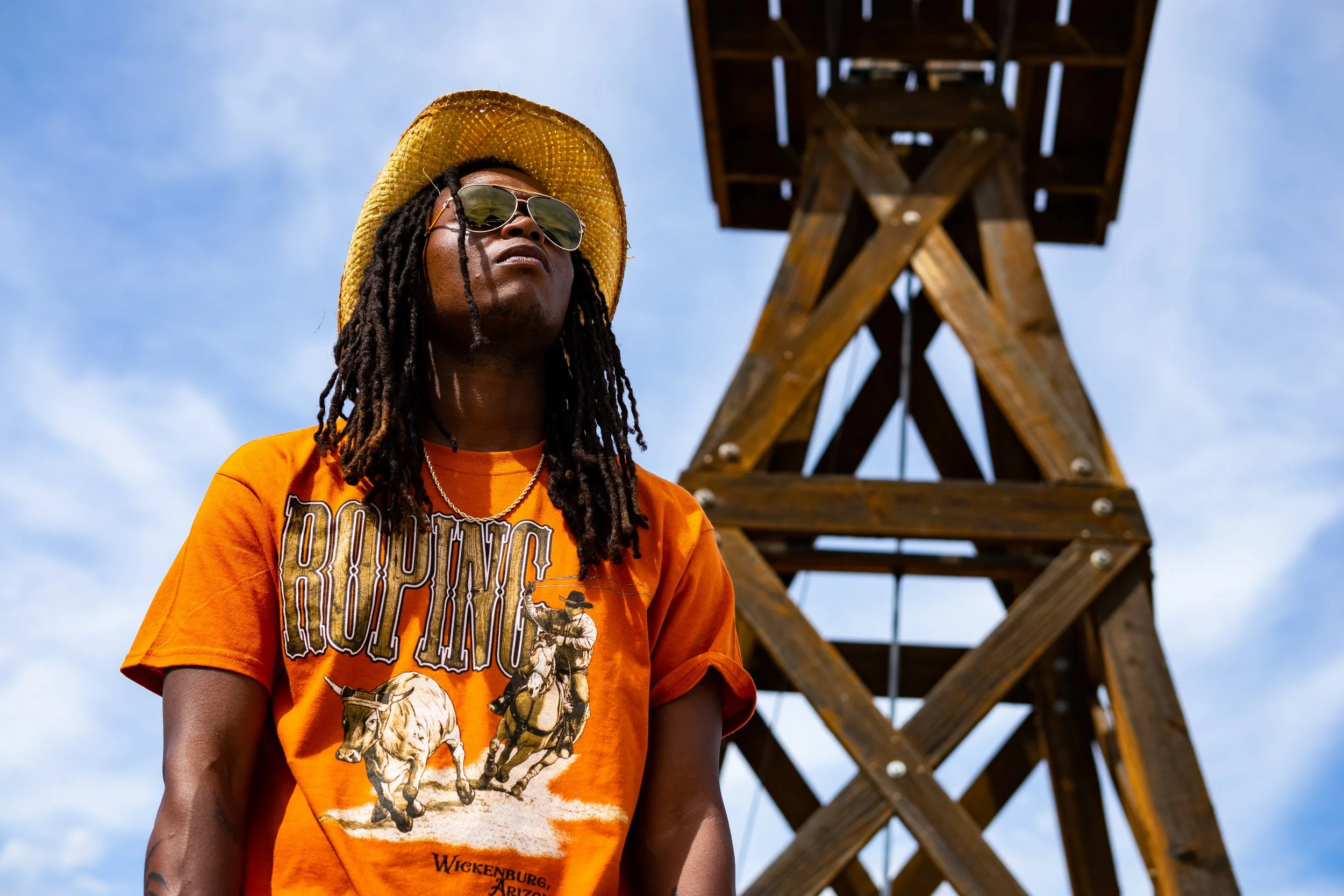 A person with long dreadlocks wearing sunglasses and a wide-brimmed straw hat, standing outdoors with a wooden tower structure in the background under a sky with scattered clouds.