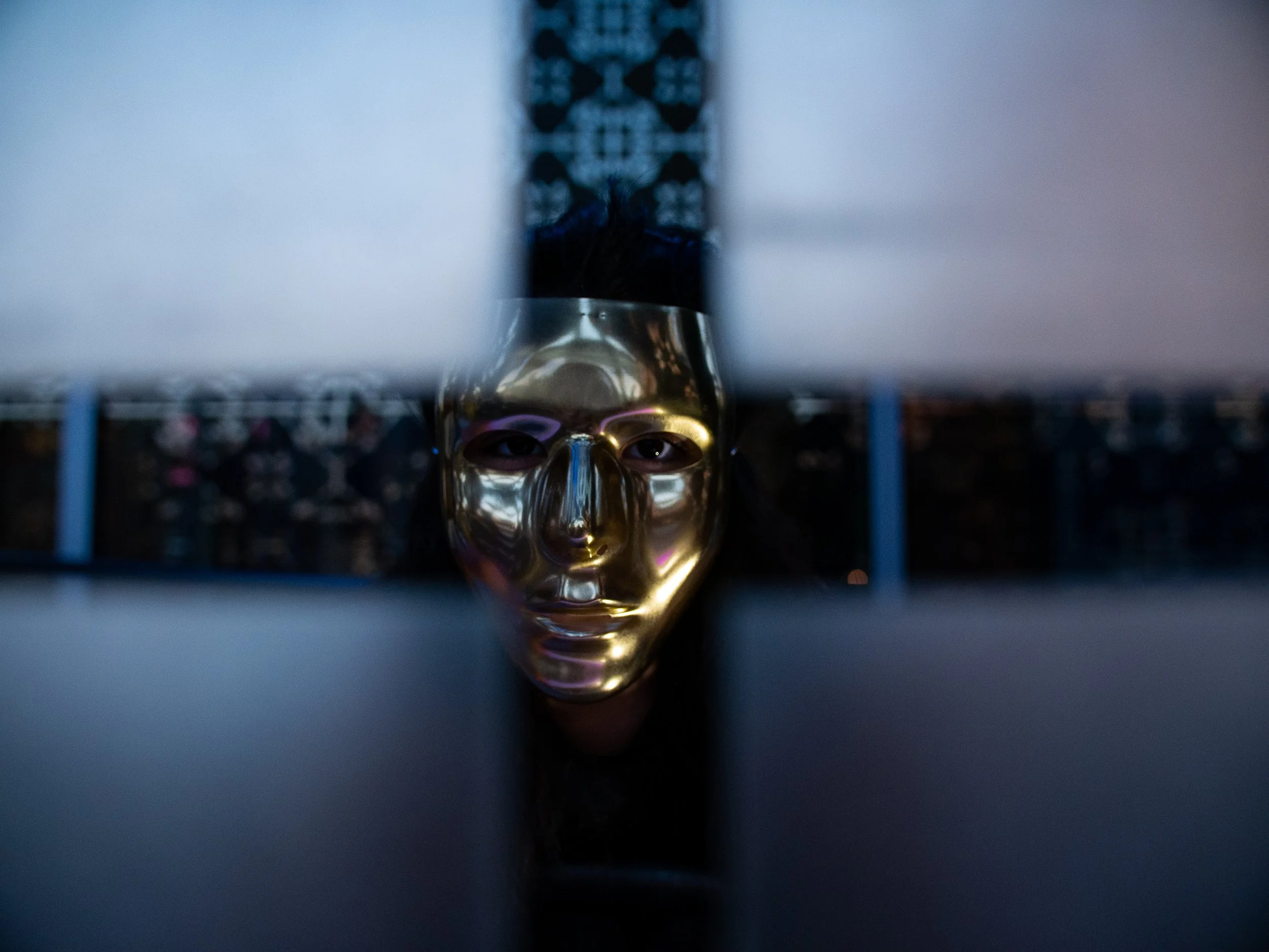 A person wears a gold-colored skull mask, peering over a dark surface with eyes visible through the eye holes.