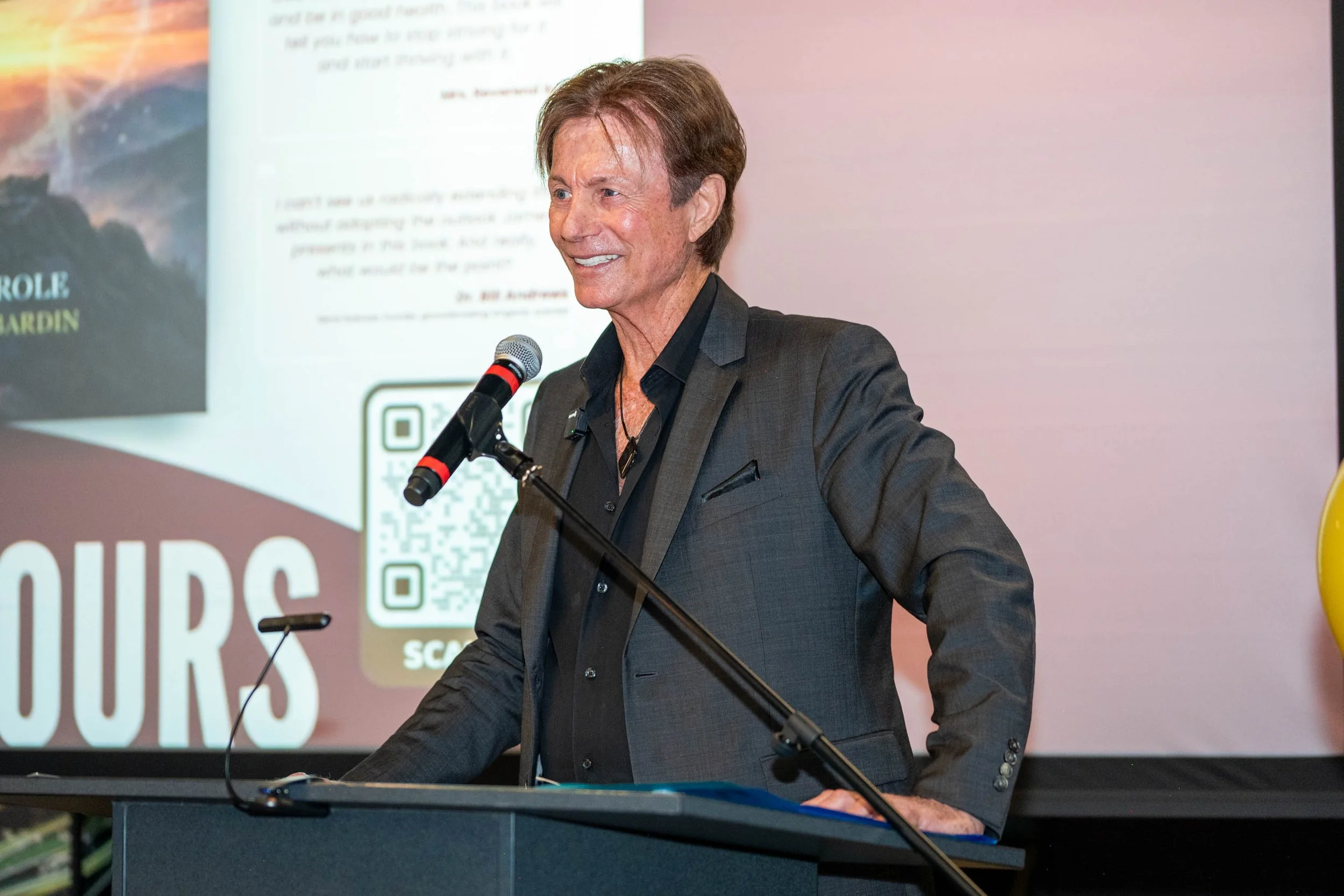 James Strole reads a chapter of his new book, The Gospel of Longevity. Wearing a dark gray jacket at a podium while smiling