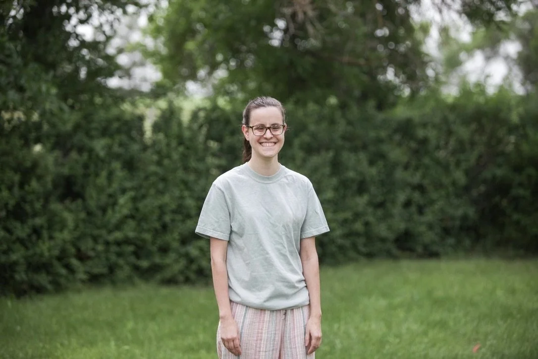 A woman with glasses, wearing a mint green t-shirt and striped pants, standing outdoors on a grassy area with green trees in the background, smiling.