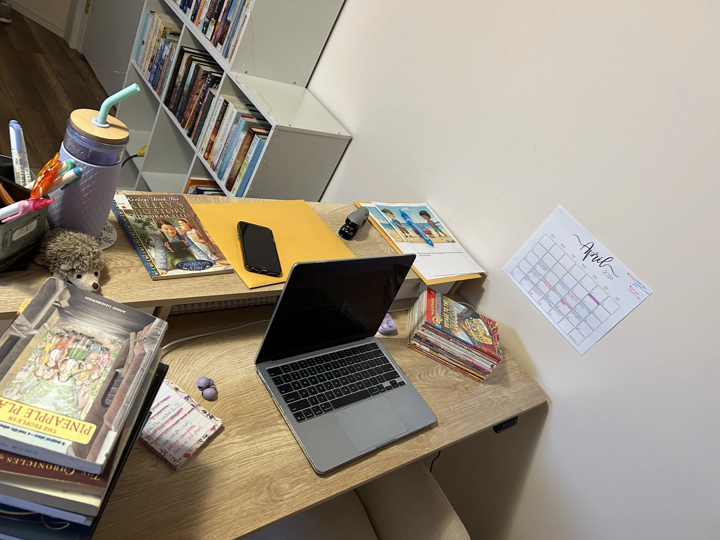 A cluttered desk with a laptop, books, a phone, notebooks, pens, and a calendar, with a white bookshelf filled with books in the background.