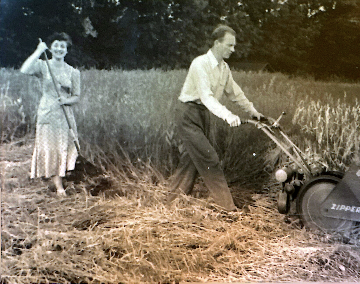 Black and white photo of a woman with a rake and a man with a lawnmower in a field.