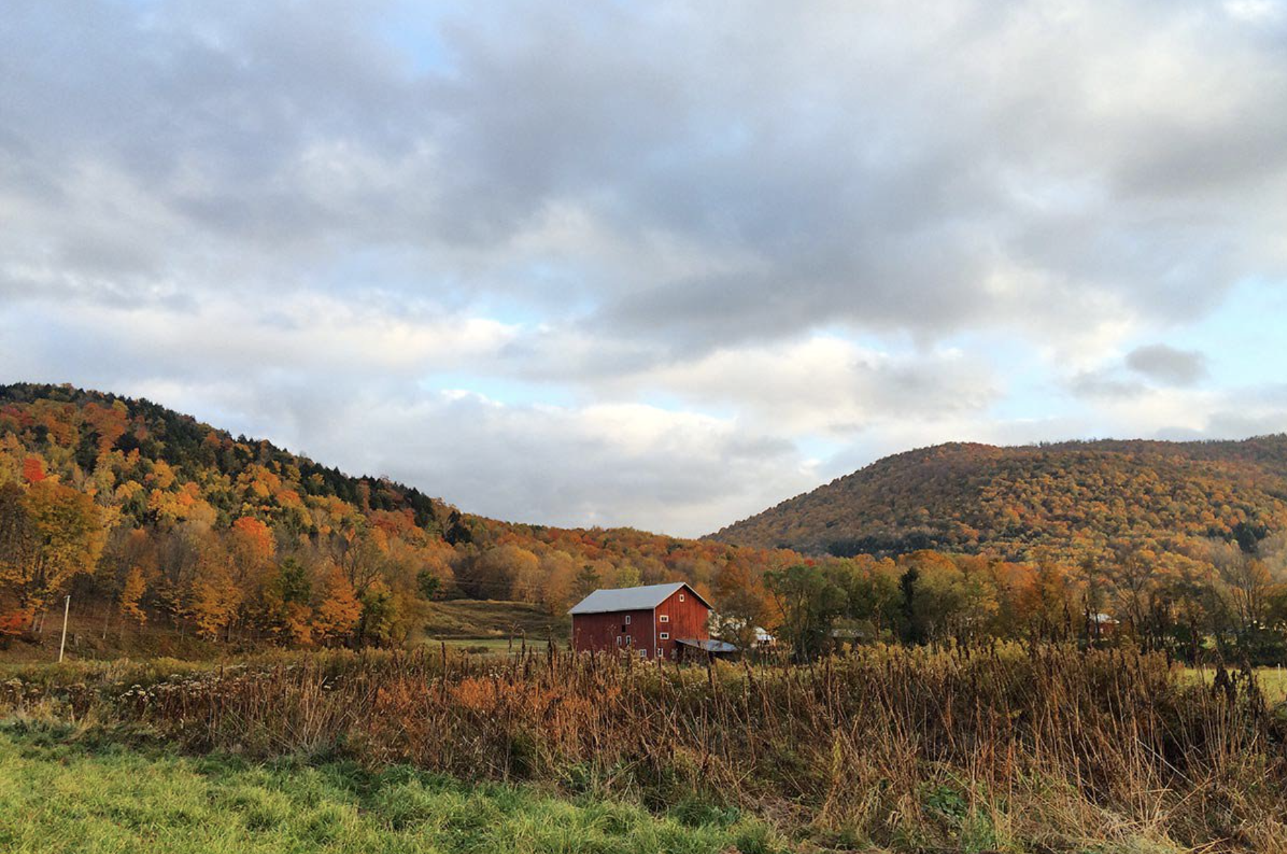 A scenic farm landscape in autumn with rolling hills covered in colorful fall foliage, a red barn, and a partly cloudy sky.