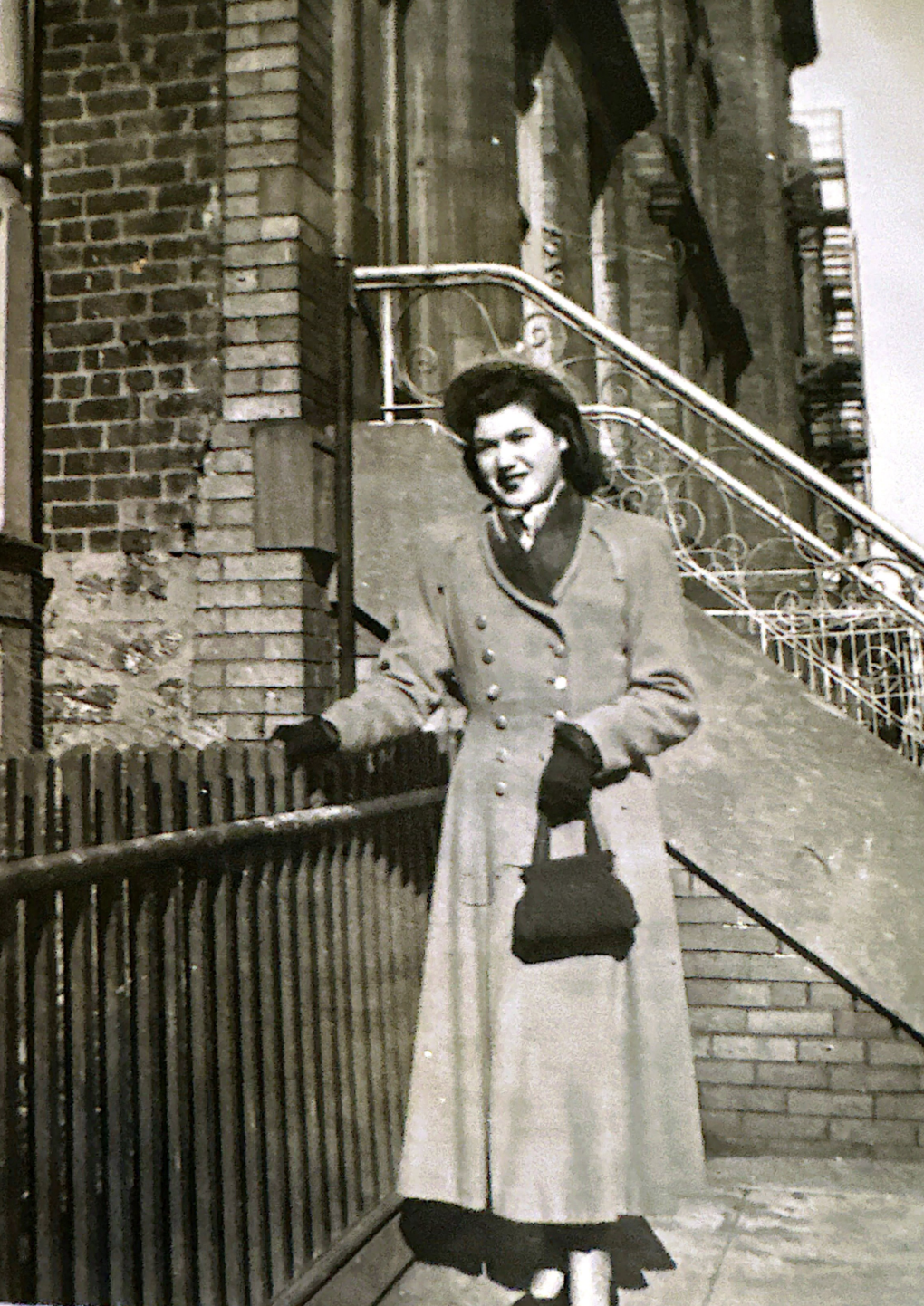 A black-and-white photo of a young woman standing on a city sidewalk near a brick building with a staircase and railing in the background. She is wearing a long coat, gloves, and holding a small bag.