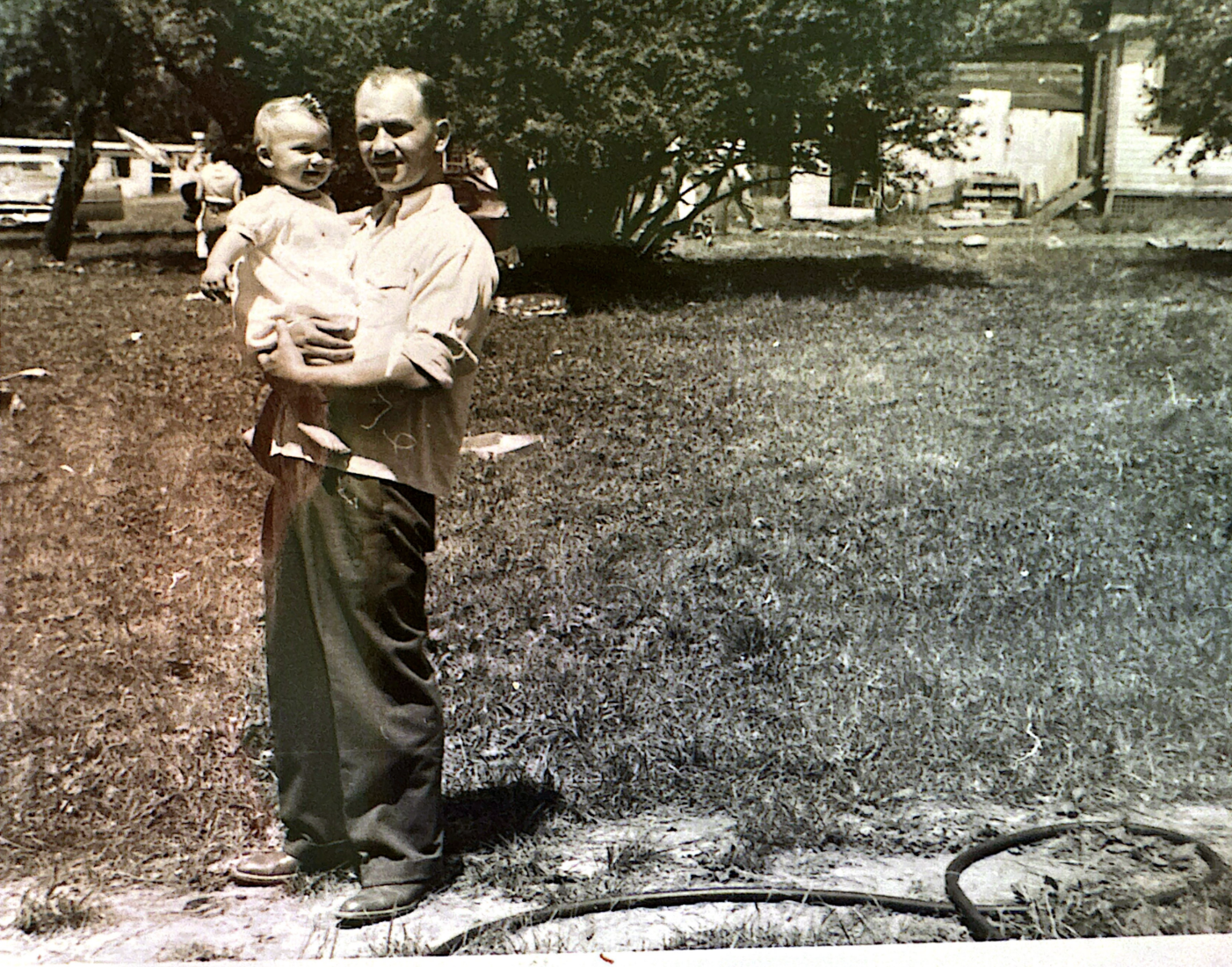 A man holding a young girl in an outdoor yard with grass, trees, and a house in the background.
