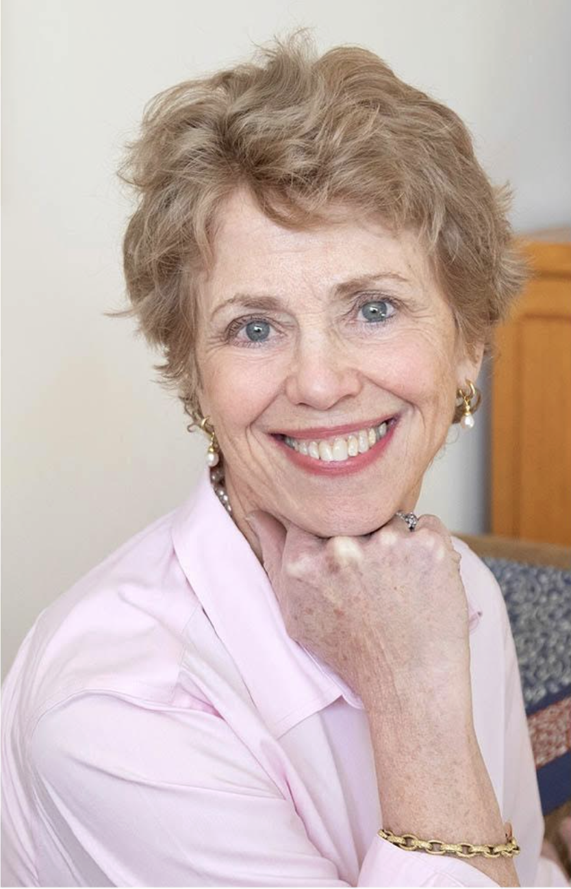 An elderly woman with short, curly blonde hair, blue eyes, and a broad smile, wearing pearl earrings, a gold bracelet, a ring, and a light pink blouse, resting her chin on her hand.