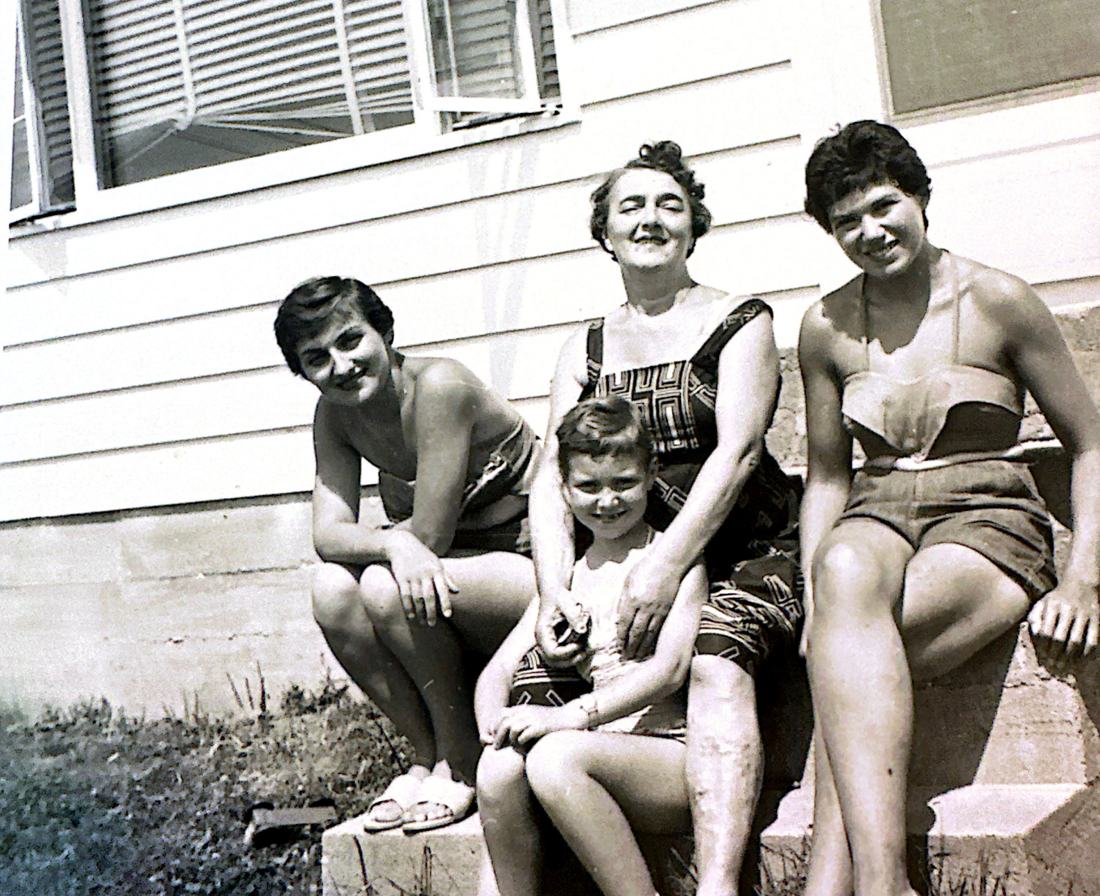 A vintage black-and-white photo of four people sitting on a concrete step outside a house, including three young women, a young boy, and a dog, with a house with a window and siding in the background.