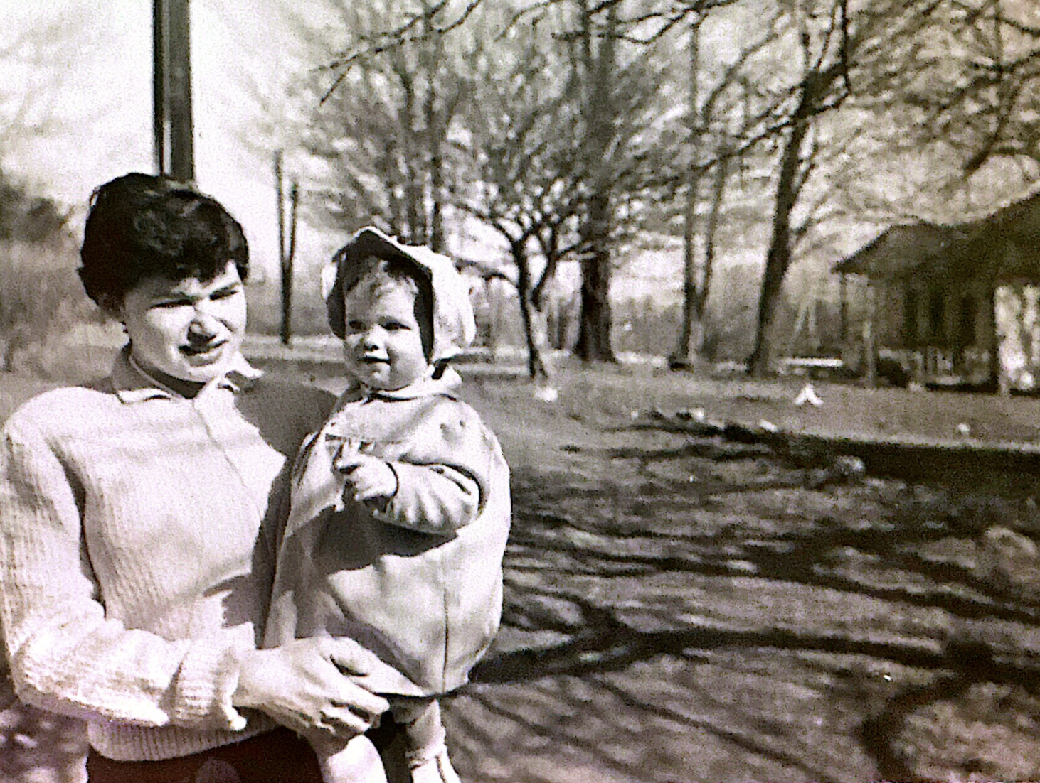 A woman holding a young girl outdoors in a park-like setting with leafless trees and a small house in the background.