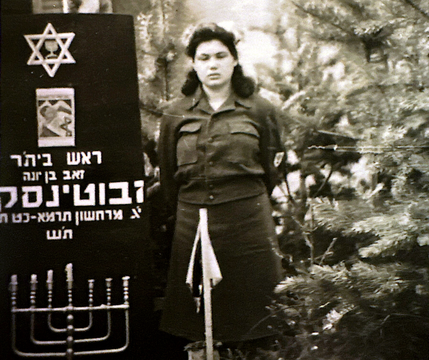 A black-and-white photo of a woman in military uniform standing outdoors. To her left, there is a blackboard with a Star of David, a menorah, and Hebrew text.