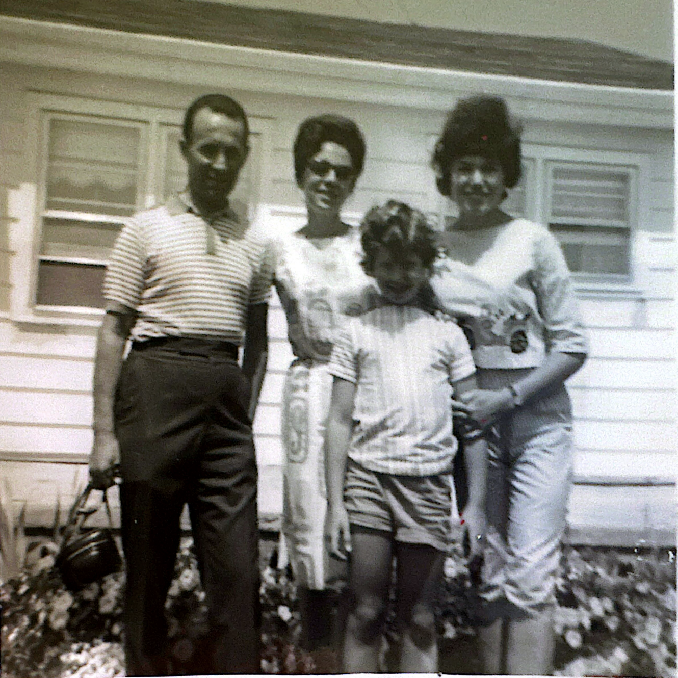 Black and white photo of four people standing outside in front of a house with horizontal siding. The group includes two women and two men, all smiling. One woman is wearing sunglasses and a short-sleeve top, another woman has a high hairstyle and is wearing a long-sleeve top. Both men are wearing collared shirts, one striped and one plain, with the man on the left holding a bag and a baseball bat. The others are holding objects, and there's a dog in front of the group.