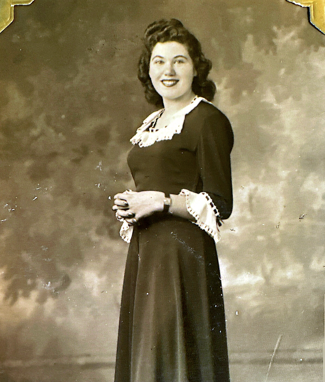 A black-and-white portrait of a woman with dark wavy hair, wearing a long dress with lace details on the sleeves and collar, standing outdoors with a blurred natural background.