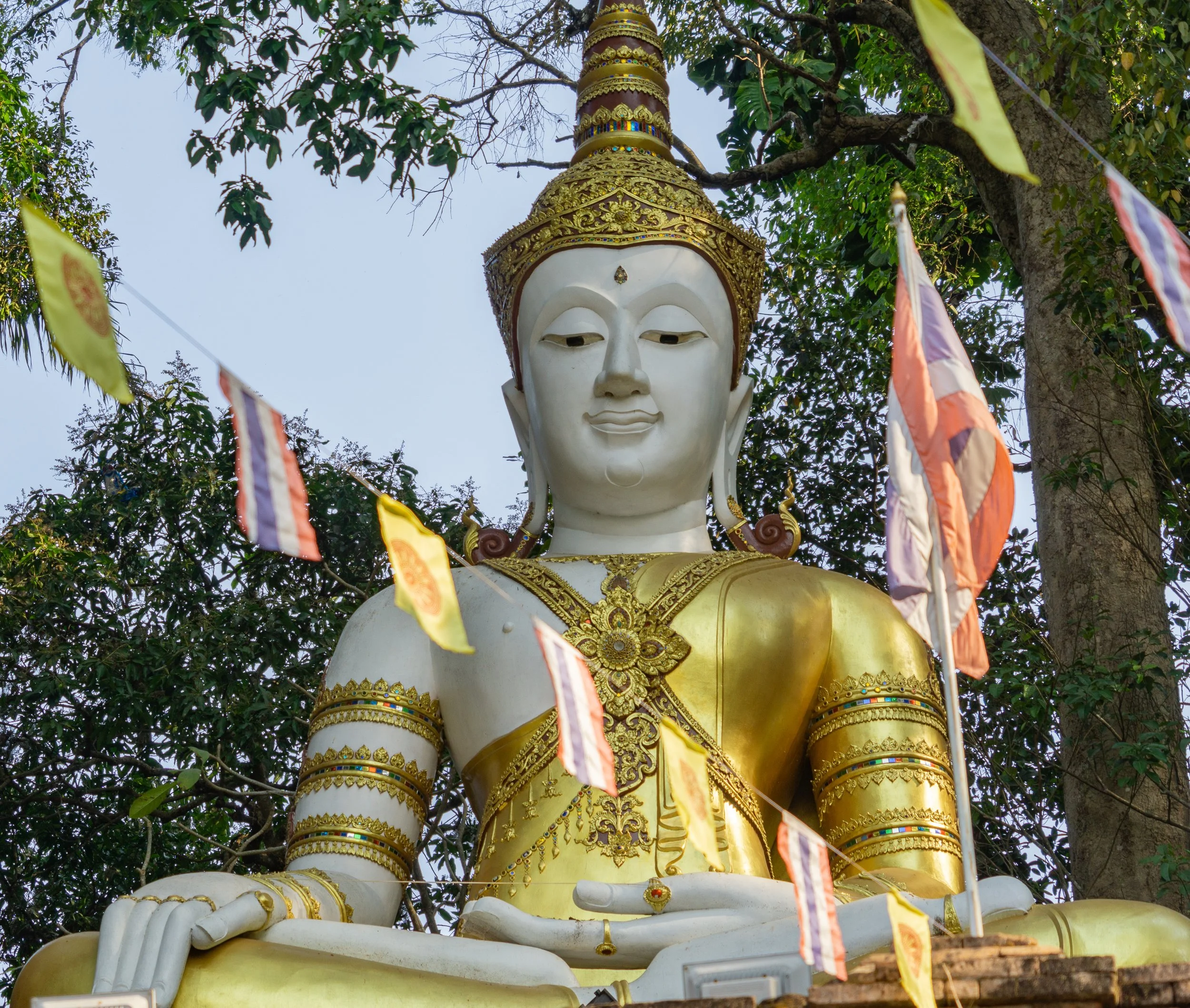 Large white Buddha statue framed by colorful prayer flags beneath trees in northern Thailand.