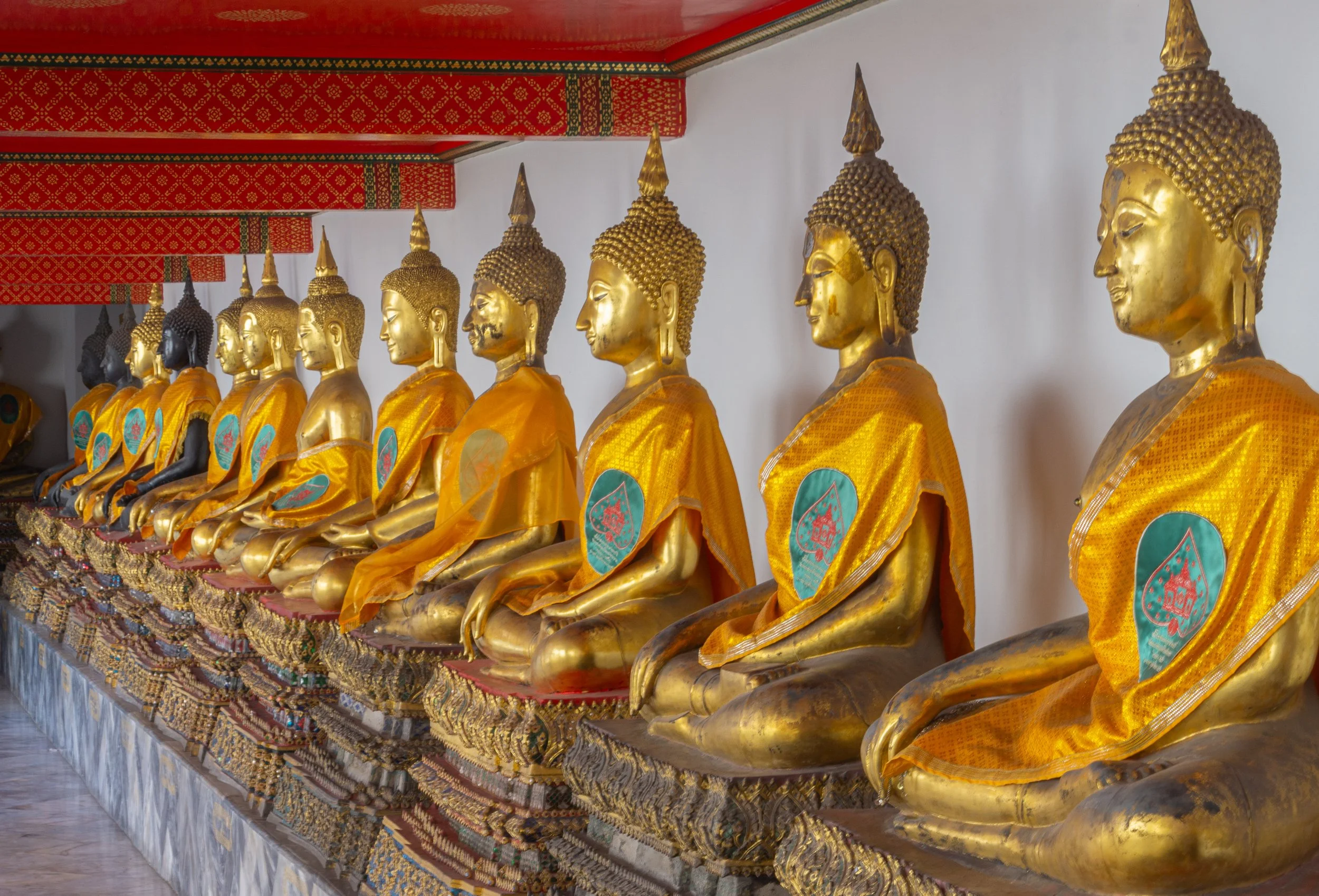 A row of golden Buddha statues draped in saffron robes lines a corridor inside Wat Pho Temple in Bangkok, Thailand.