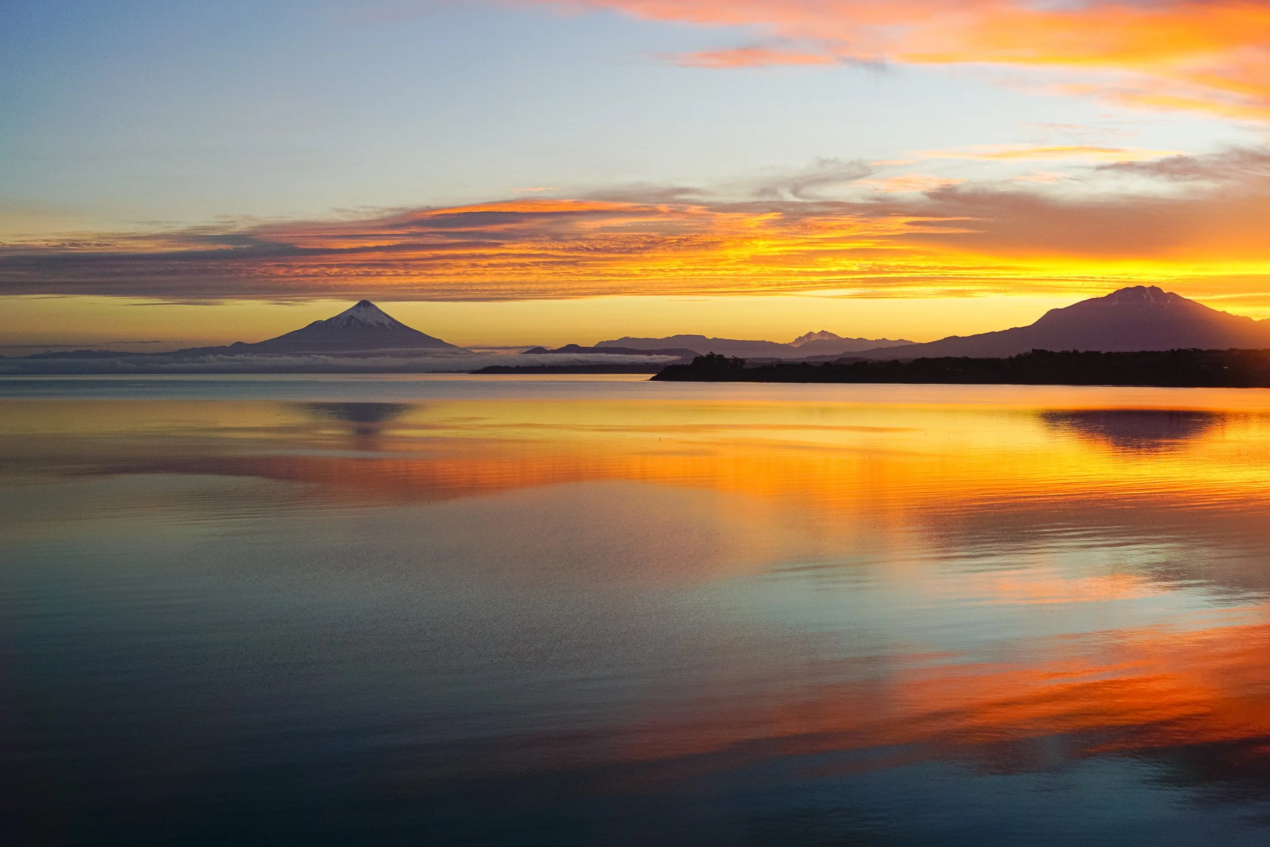 Brilliant orange sunset reflected across Lake Llanquihue in Chile, with distant mountains silhouetted along the horizon.