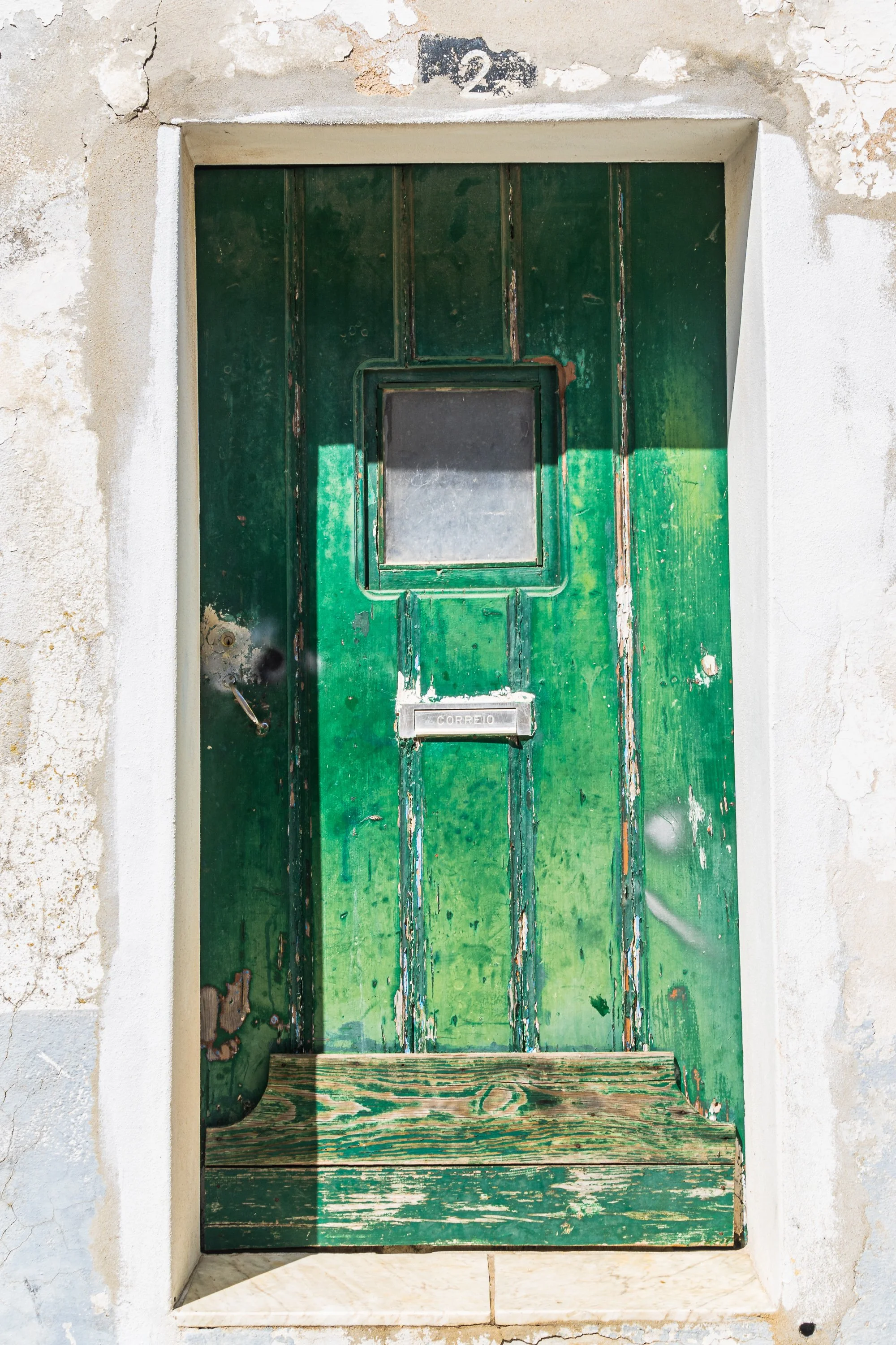 Weathered green wooden door with peeling paint, small square window, and mail slot set within a whitewashed stone doorway in Portugal.