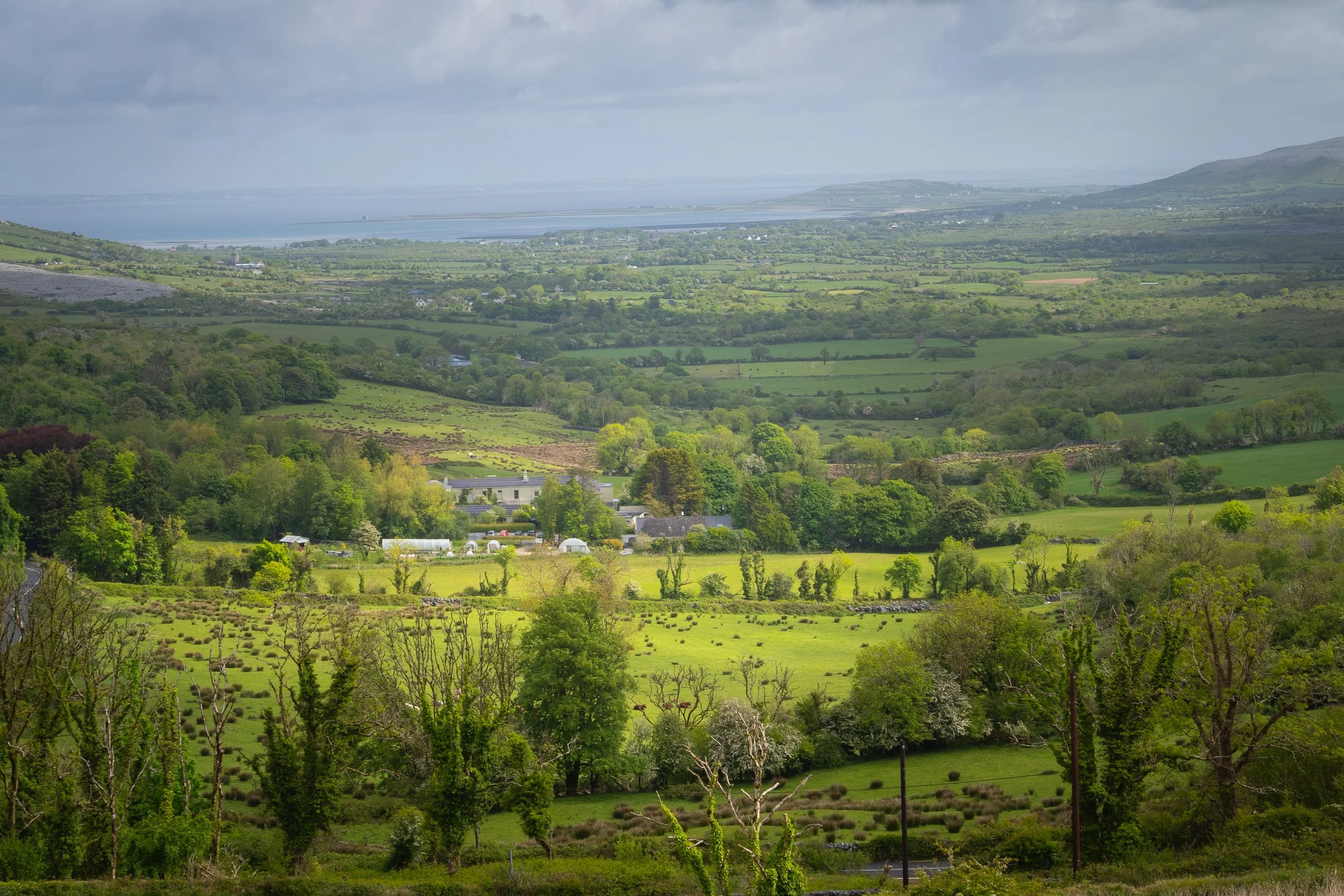 Panoramic view of the Irish countryside with patchwork green fields, rolling hills, scattered farms, and a distant coastline beneath dramatic spring clouds.