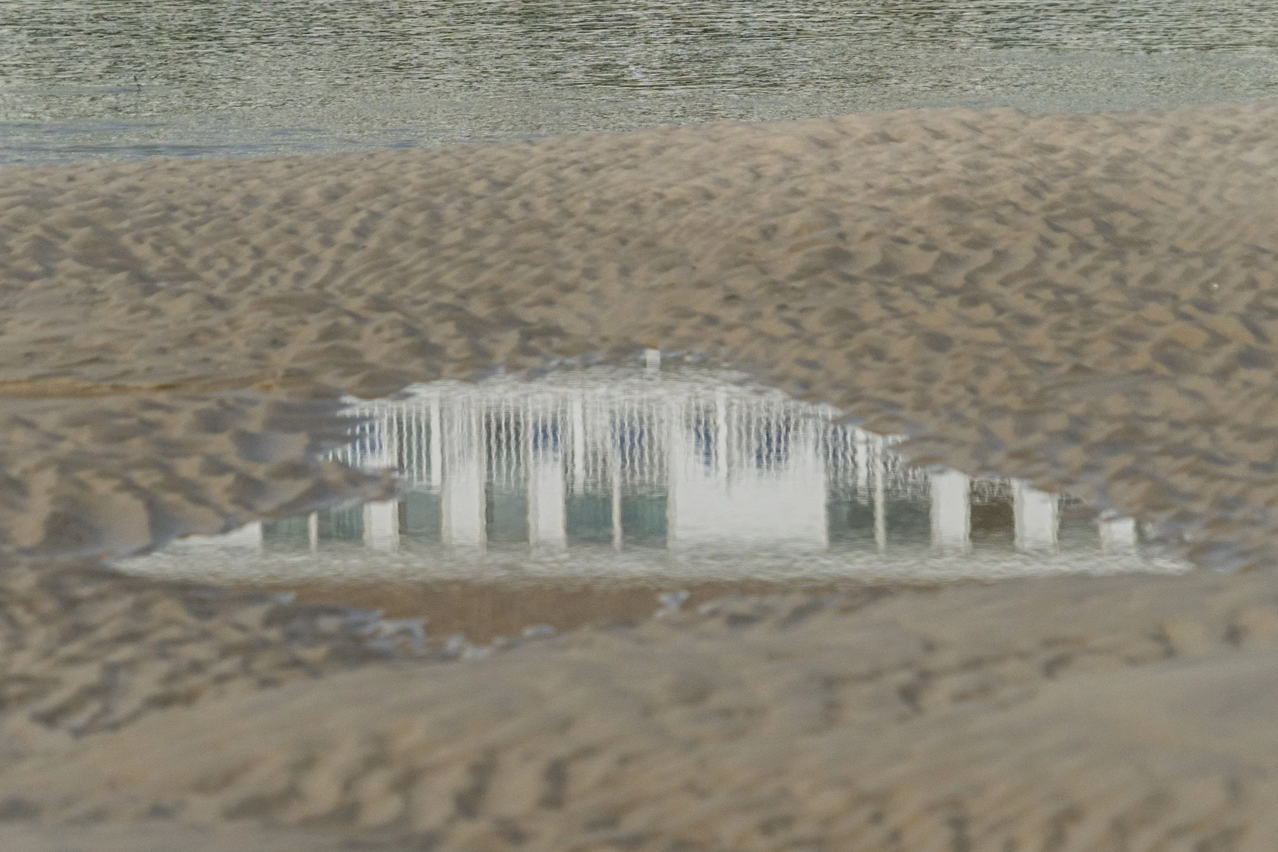 Reflection of a white beachfront building appearing in a shallow pool of water surrounded by rippled sand and footprints on Ogunquit Beach in Maine.