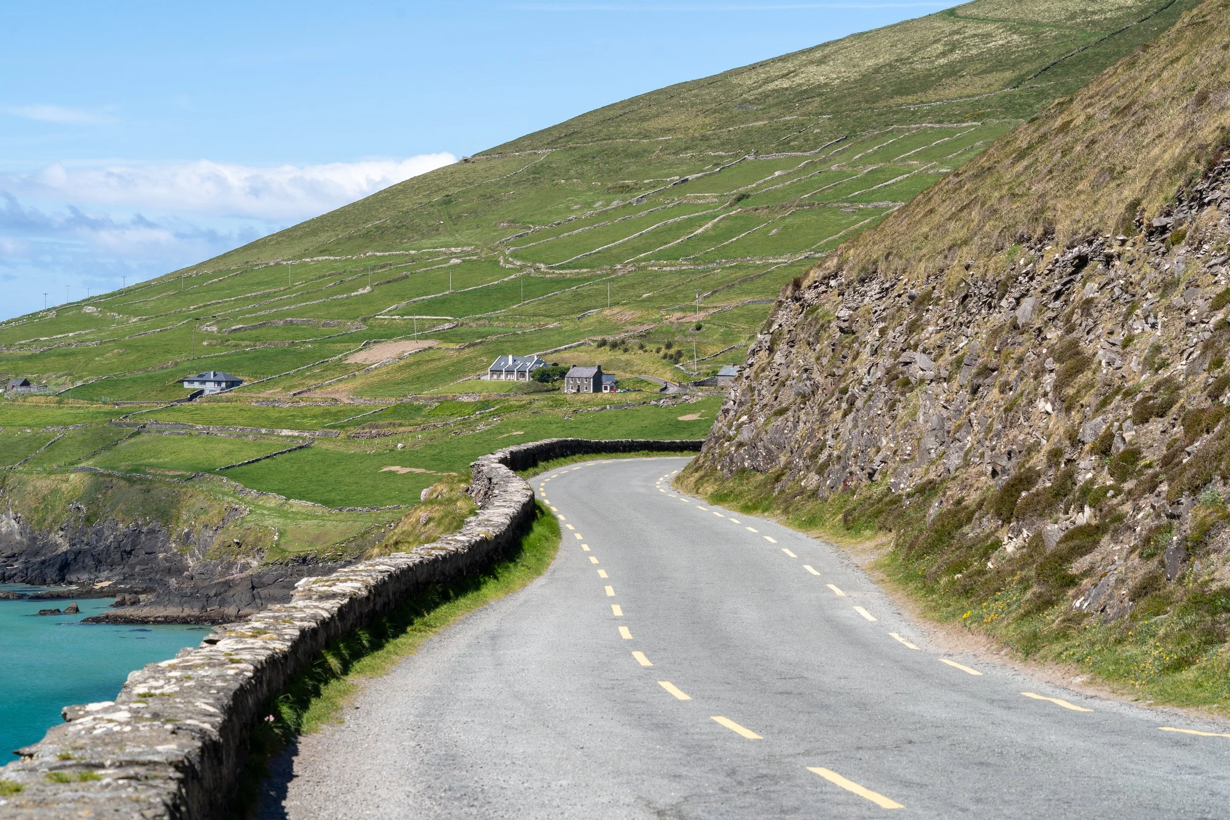 A winding coastal road follows the rugged cliffs of the Dingle Peninsula along Ireland's Wild Atlantic Way, bordered by ancient stone walls and emerald-green hillsides.