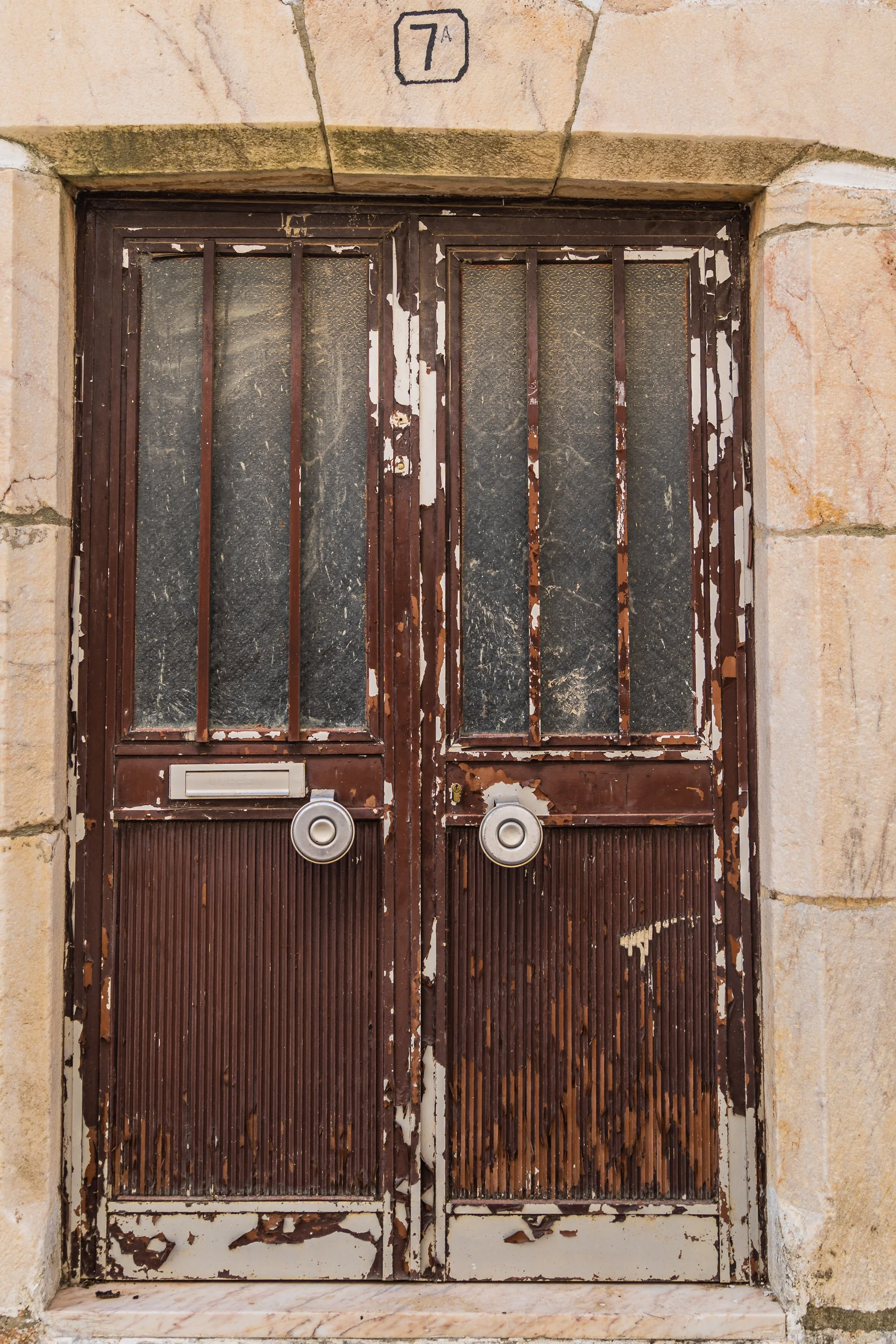 A weathered brown double door in Portugal with peeling paint, ribbed wood panels, frosted glass, and circular metal door pulls framed by aged limestone.