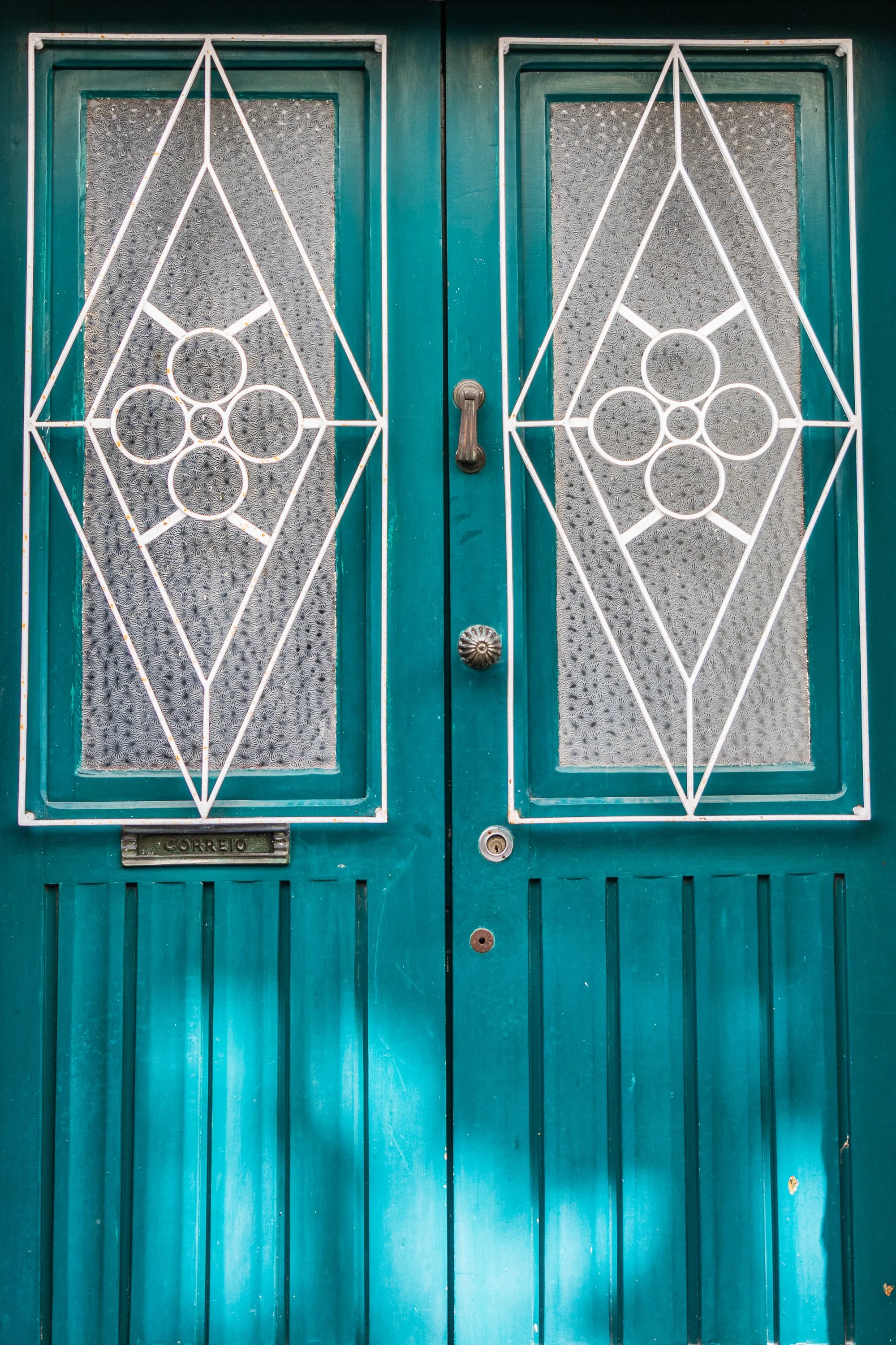 Teal double doors with decorative white wrought-iron geometric panels and textured glass in a historic Portuguese building.