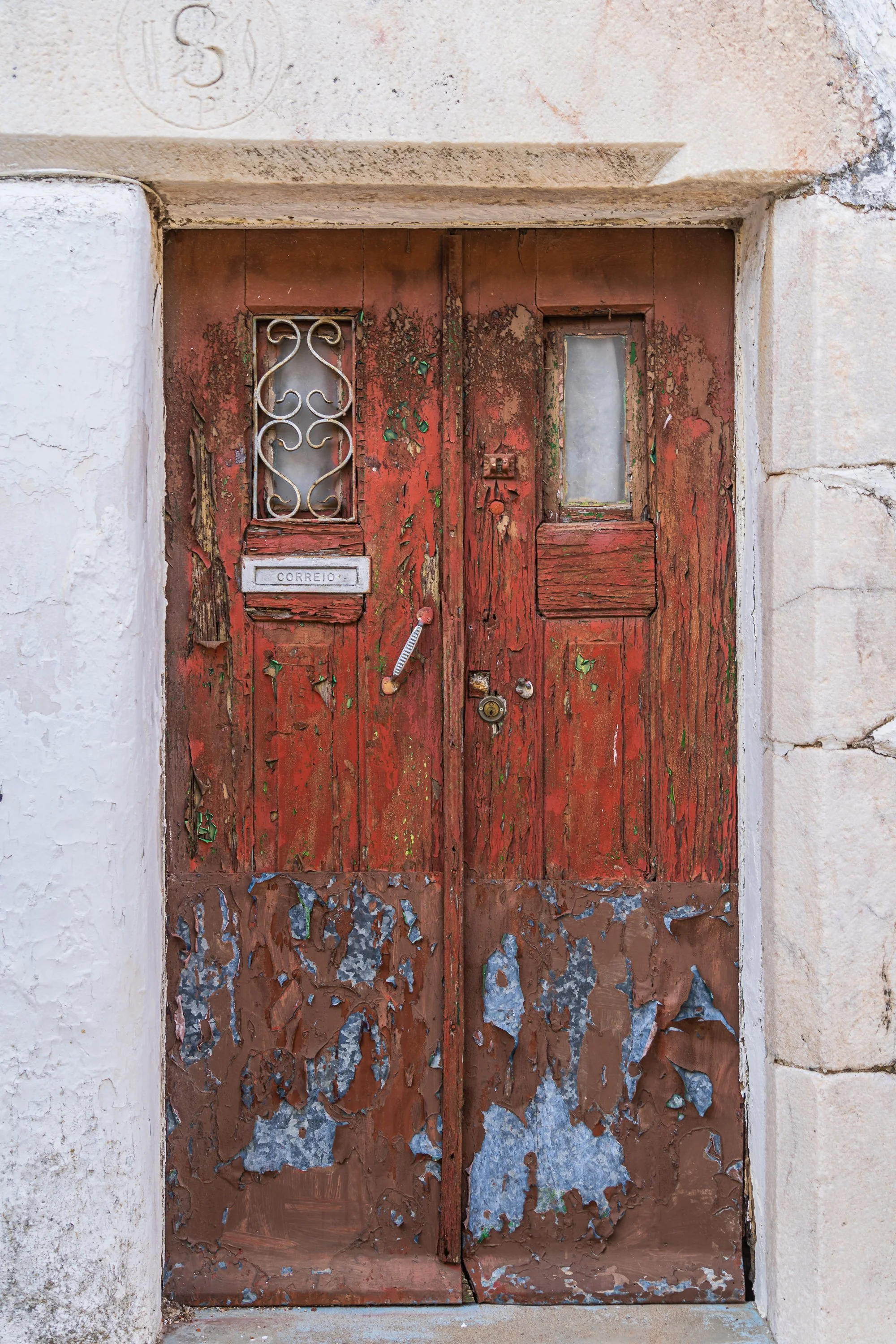 Heavily weathered red wooden door in Portugal with peeling paint, antique mail slot, decorative wrought iron window grille, and centuries of layered texture.