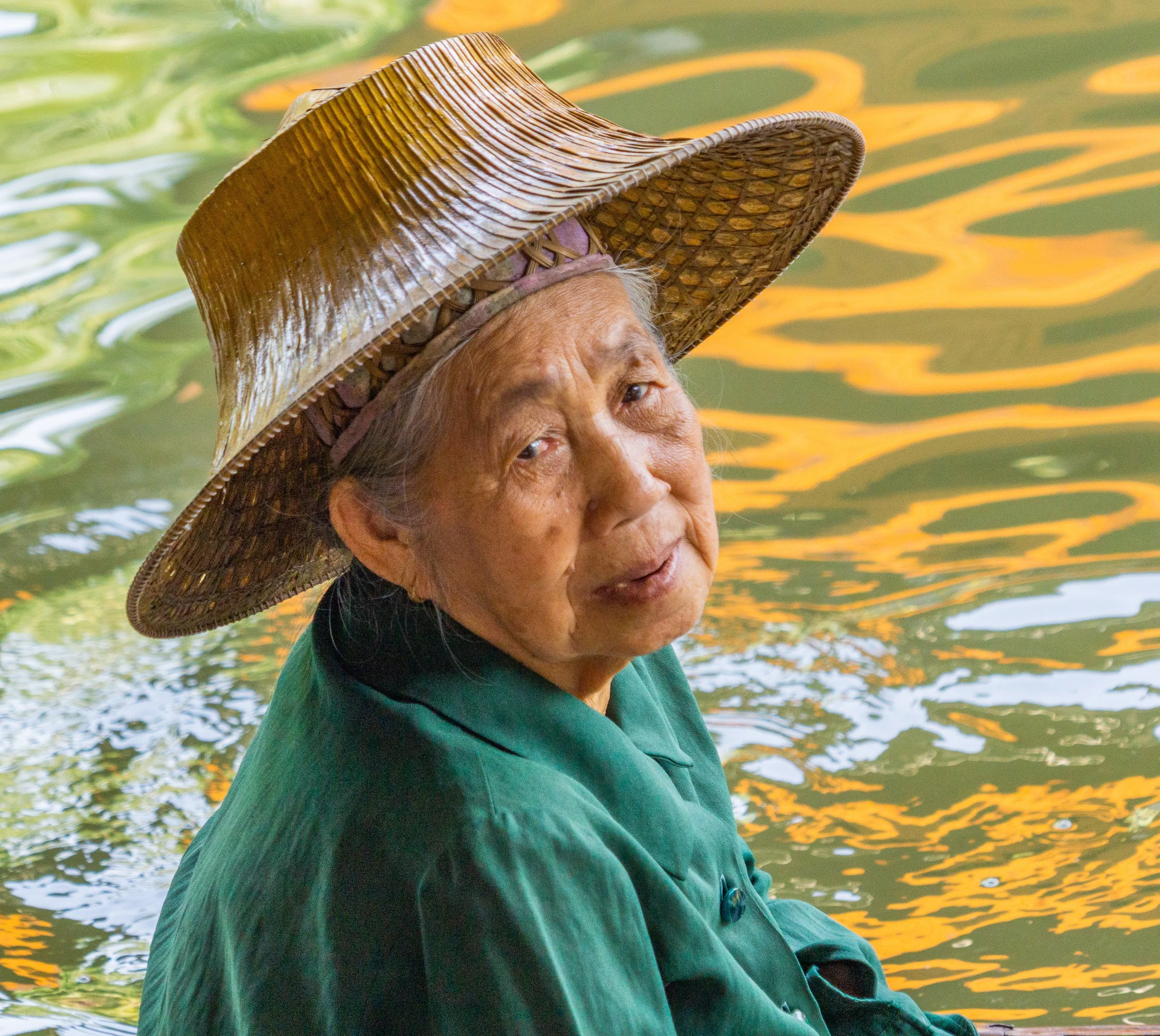 Elderly woman in a green jacket and woven hat glances back while seated beside a canal in Thailand.