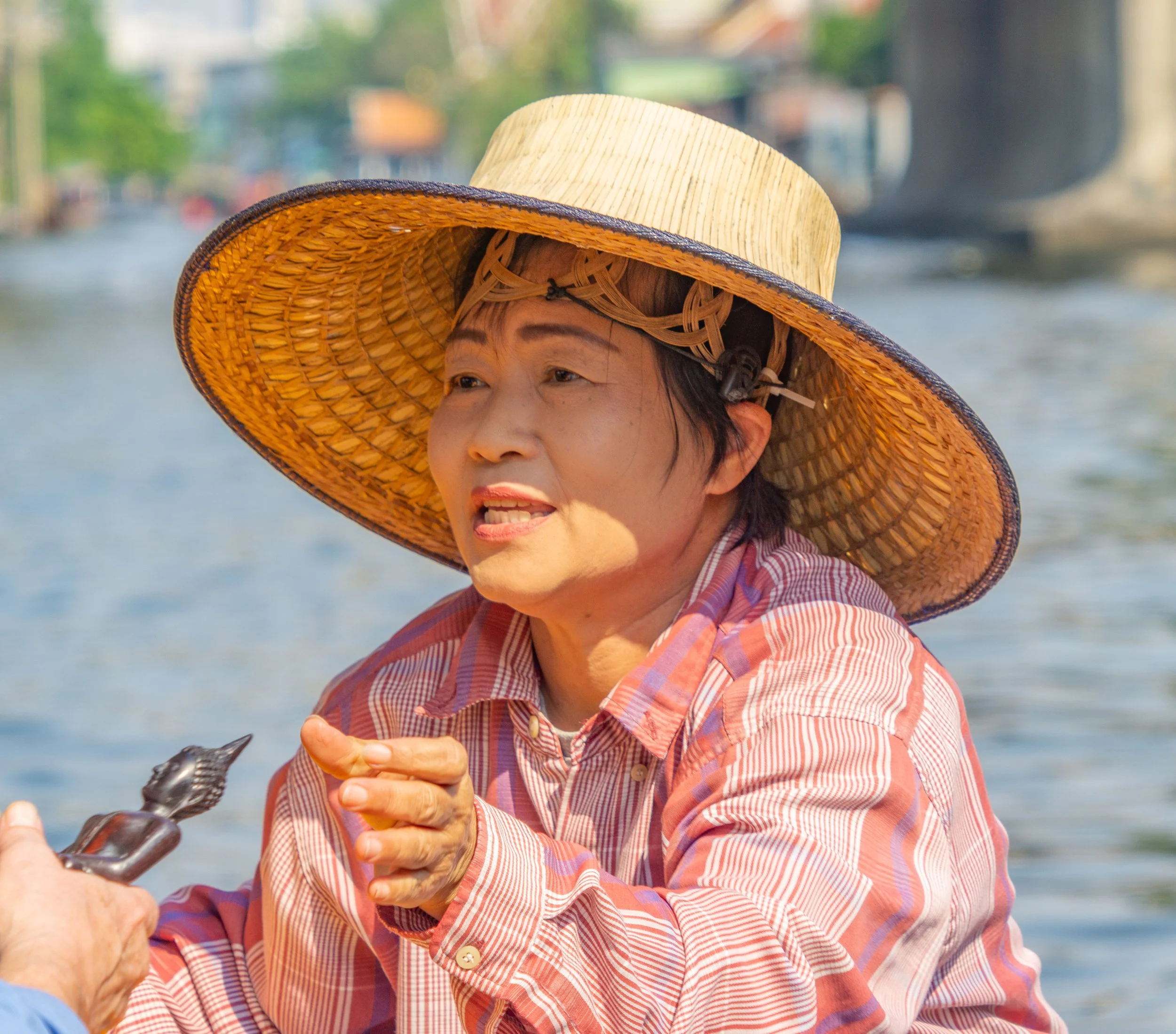Woman wearing a woven hat speaks with visitors from her boat along a canal in Bangkok, Thailand.