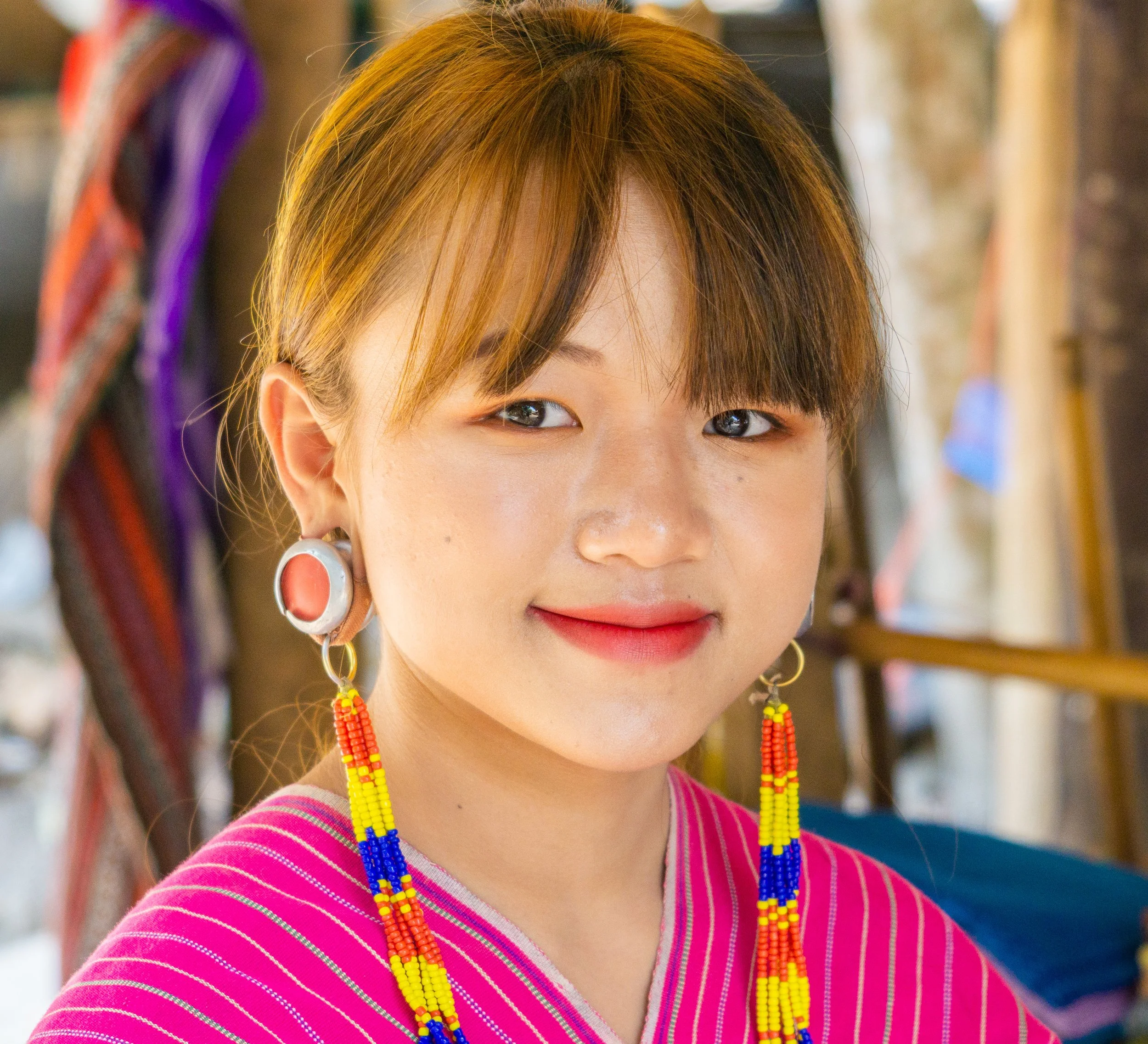 Portrait of a young Thai woman wearing traditional pink attire and colorful beaded earrings, representing the younger generation and Thailand's living cultural heritage.