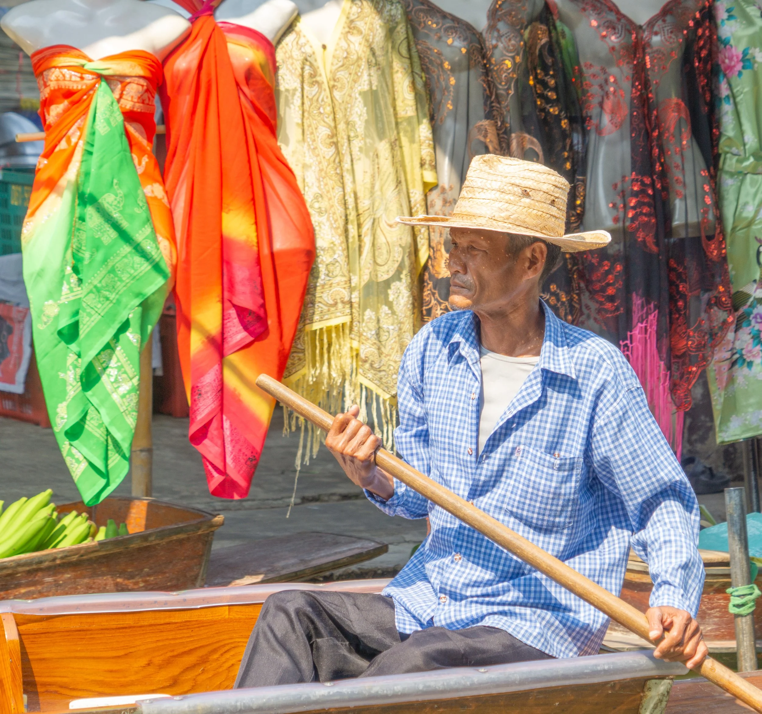 Boatman rowing a wooden boat past colorful fabrics at a floating market in Thailand.