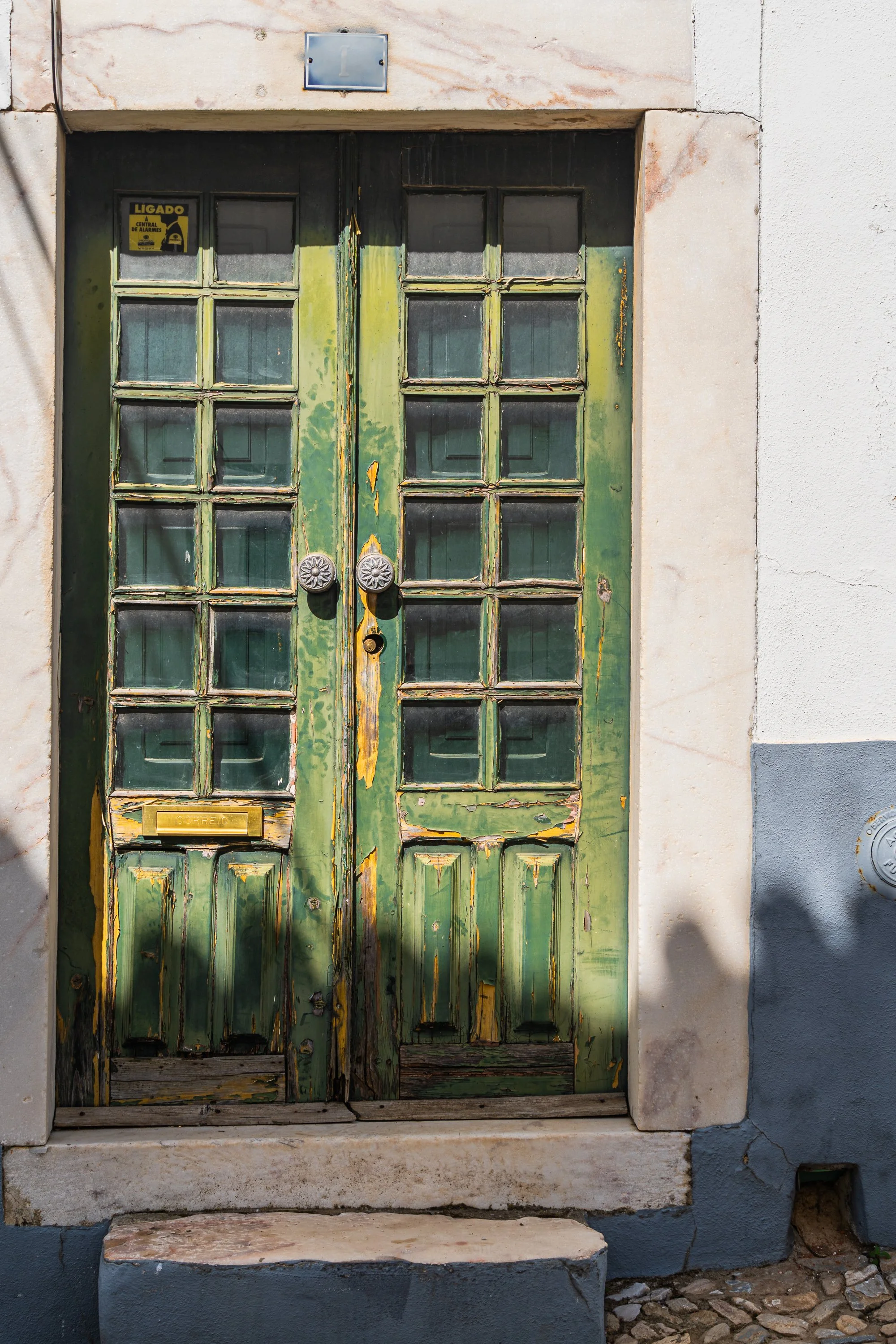 Weathered green double doors with divided glass panes, peeling paint, brass mail slot, and decorative door knobs along a historic Portuguese street.
