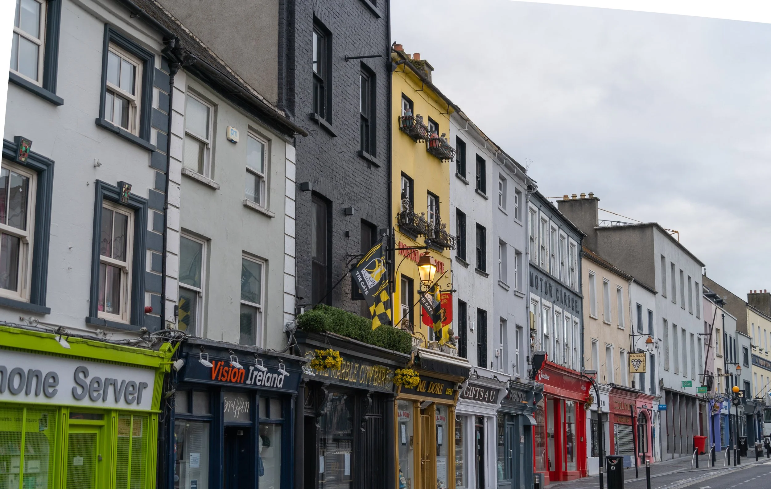 Traditional colorful pubs and storefronts lining a quiet street in Kilkenny, Ireland beneath an overcast sky.