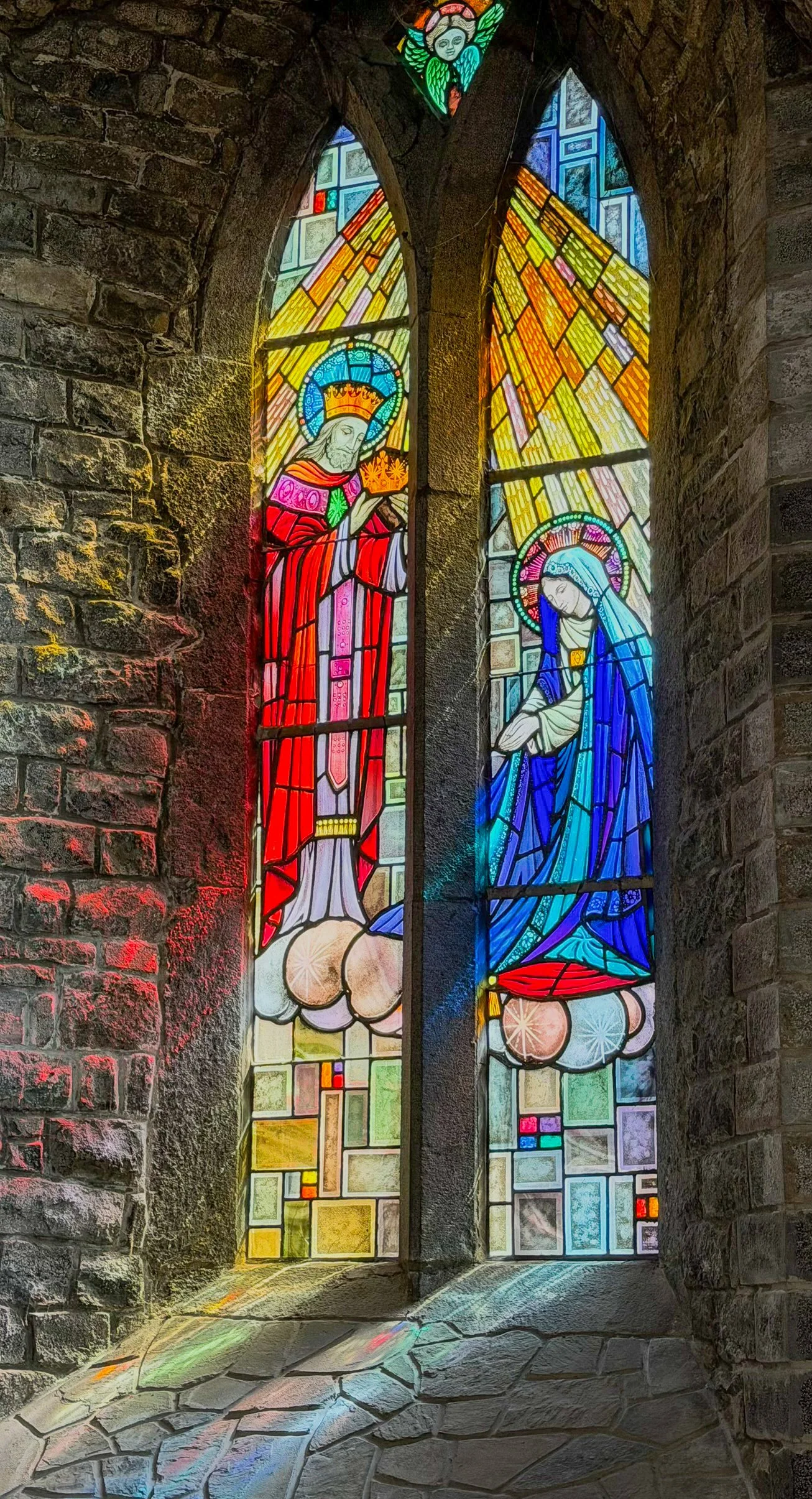 Sunlight streams through colorful stained glass windows inside Trinitarian Monastery in Adare, Ireland, casting vibrant reflections across ancient stone walls and floor.