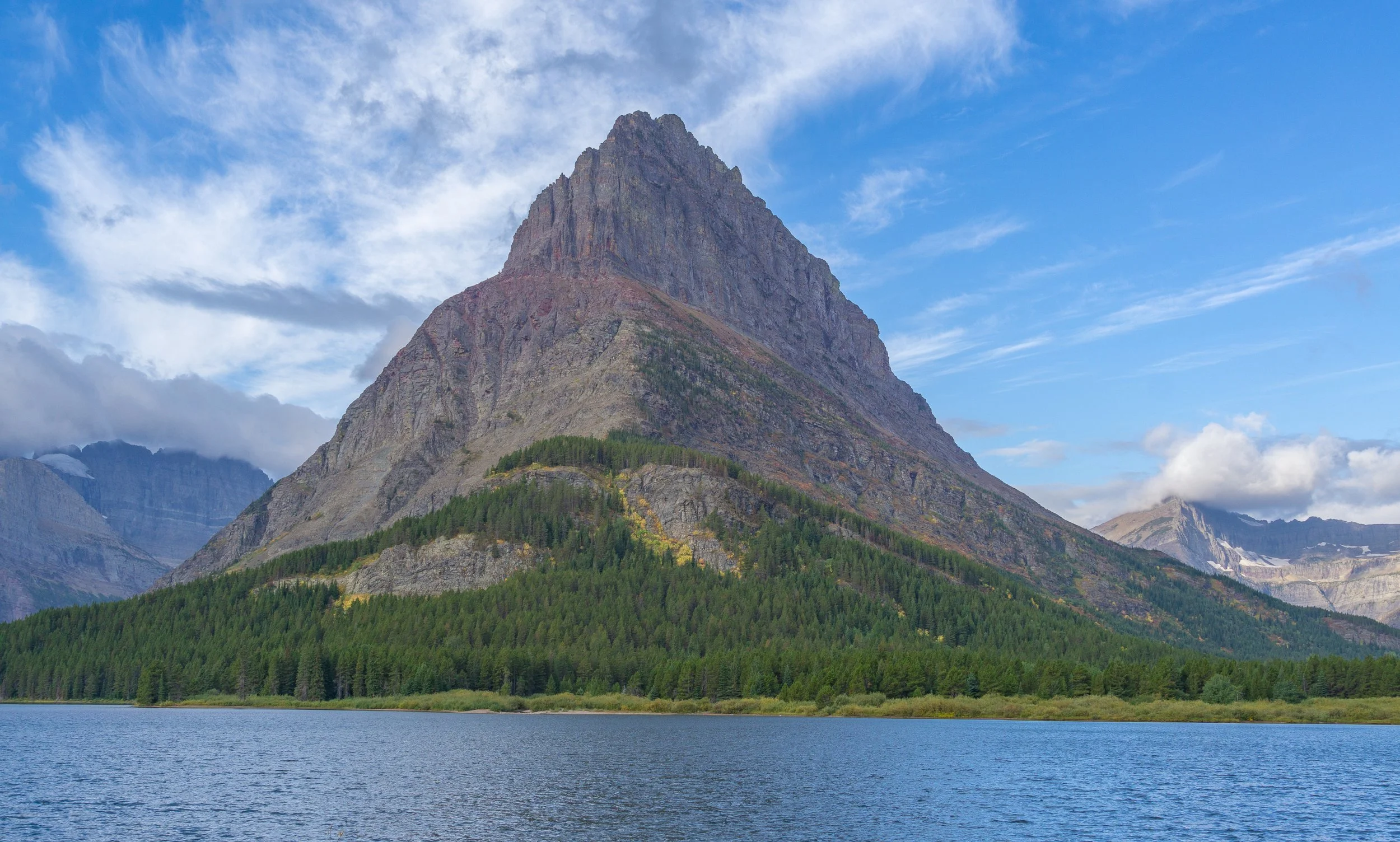 Towering mountain reflected above a tranquil lake beneath dramatic blue skies and wispy clouds in Glacier National Park, Montana.