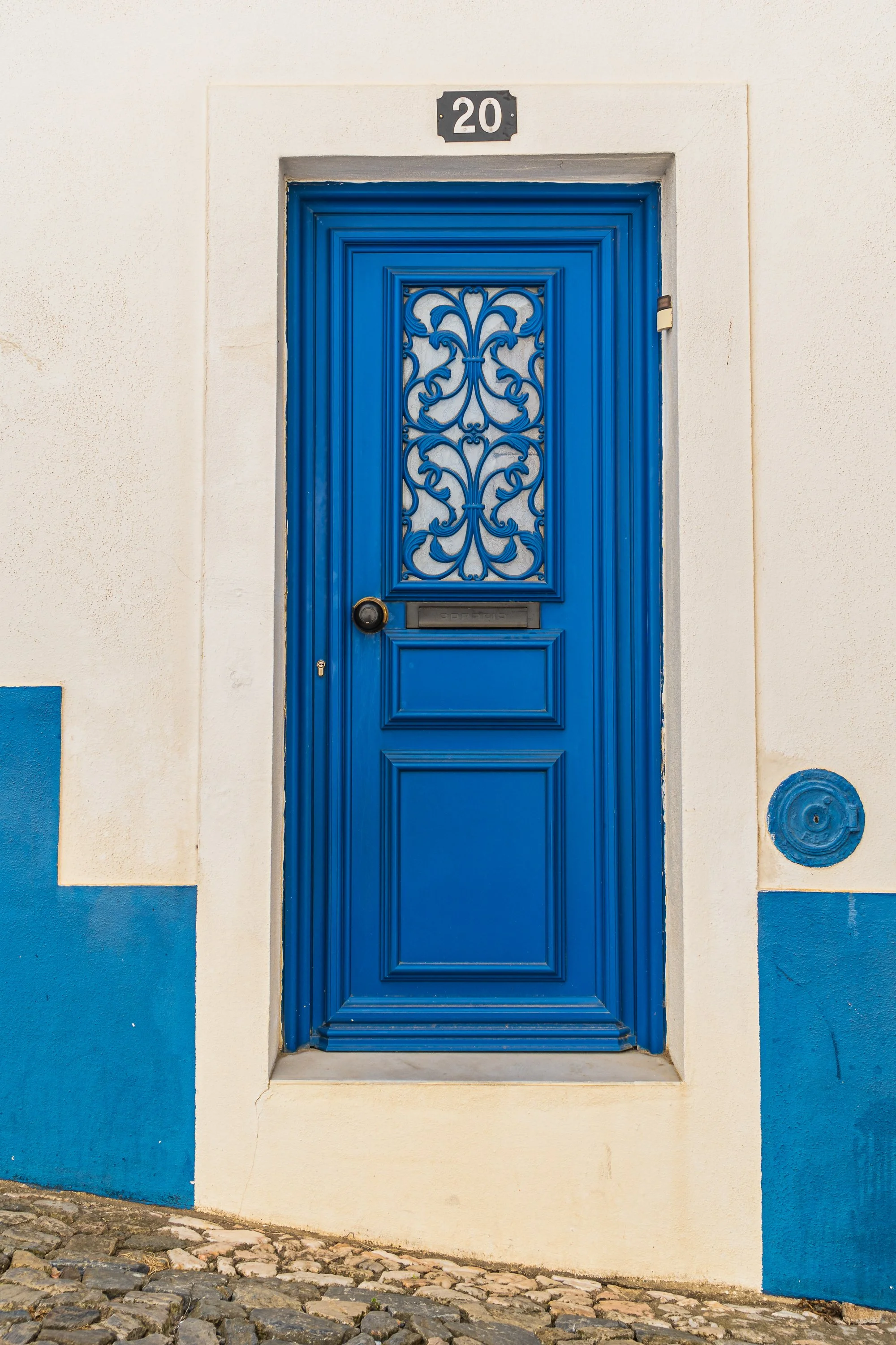 Bright cobalt blue wooden door with ornate wrought-iron grille set within a whitewashed façade and blue trim along a cobblestone street in Portugal.