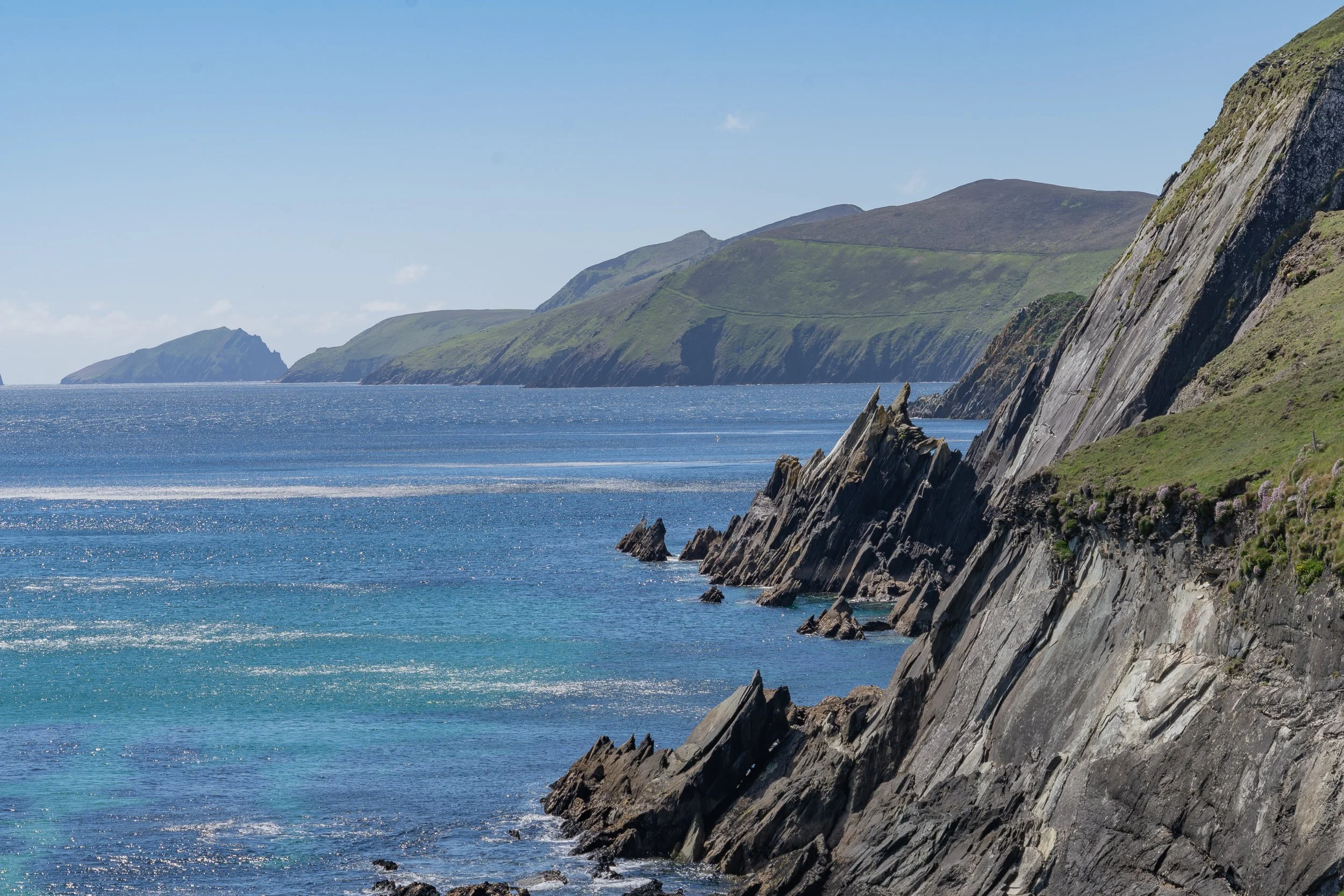 Jagged rock cliffs descend into the blue Atlantic Ocean along the Dingle Peninsula on Ireland's Wild Atlantic Way, with distant green mountains under a clear sky.
