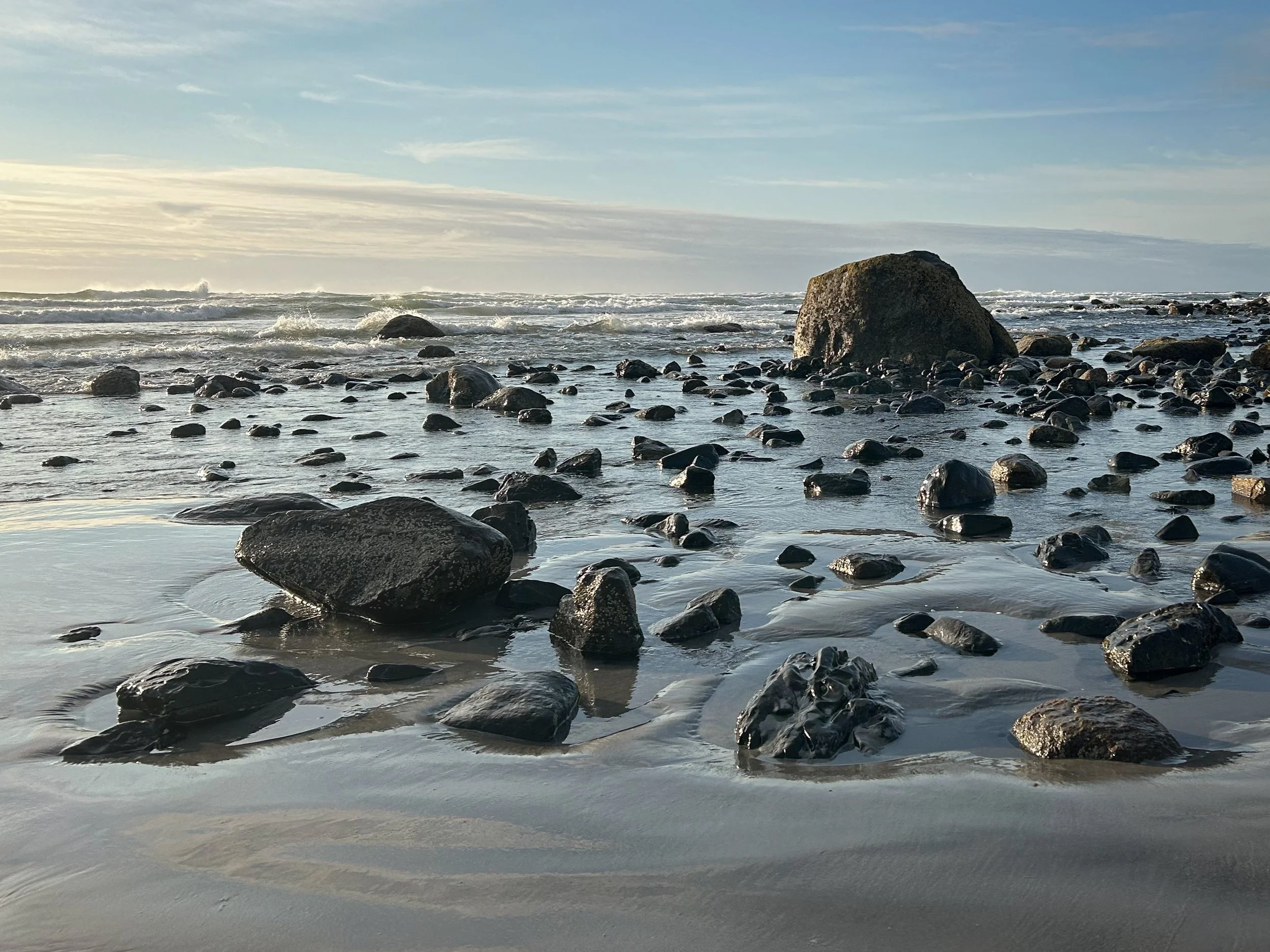 Sunlight illuminates a rocky shoreline at low tide as gentle waves wash around scattered boulders beneath a soft morning sky.