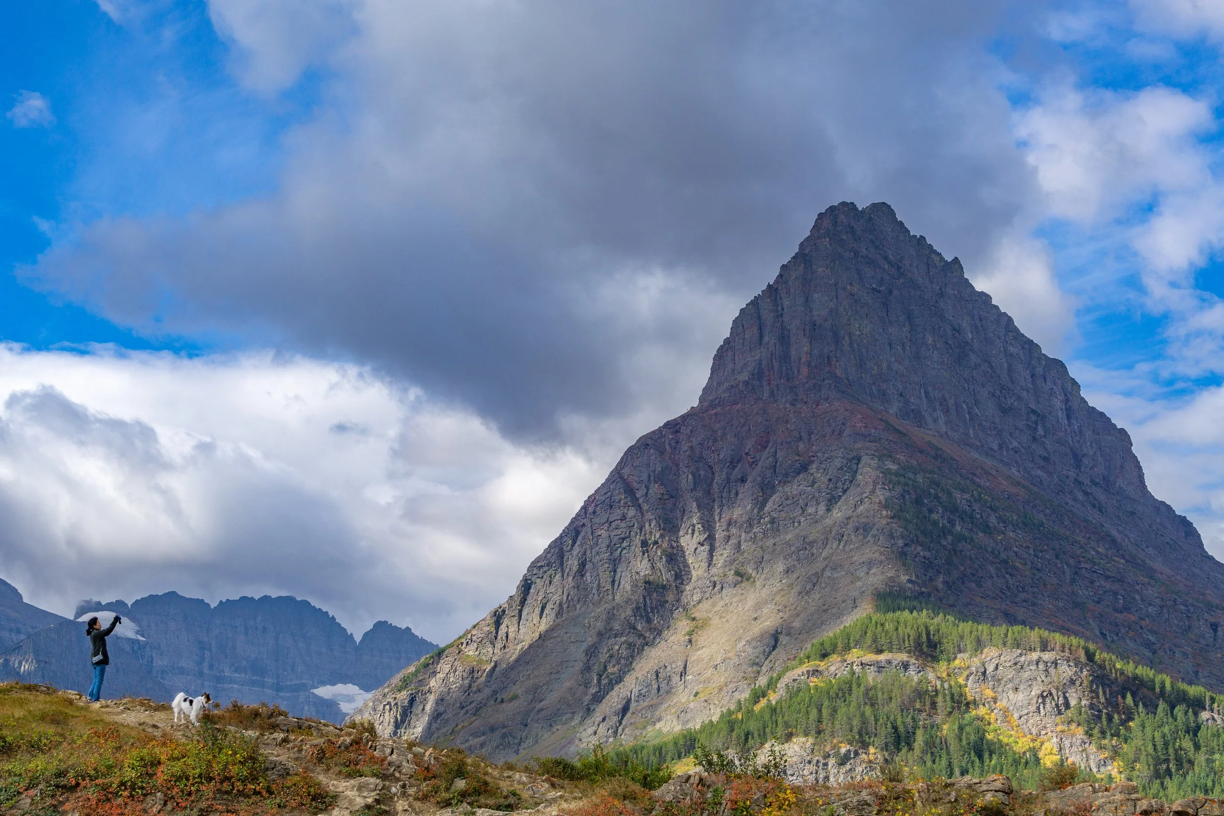 A hiker with a dog photographs a towering mountain beneath dramatic clouds in Glacier National Park, Montana.