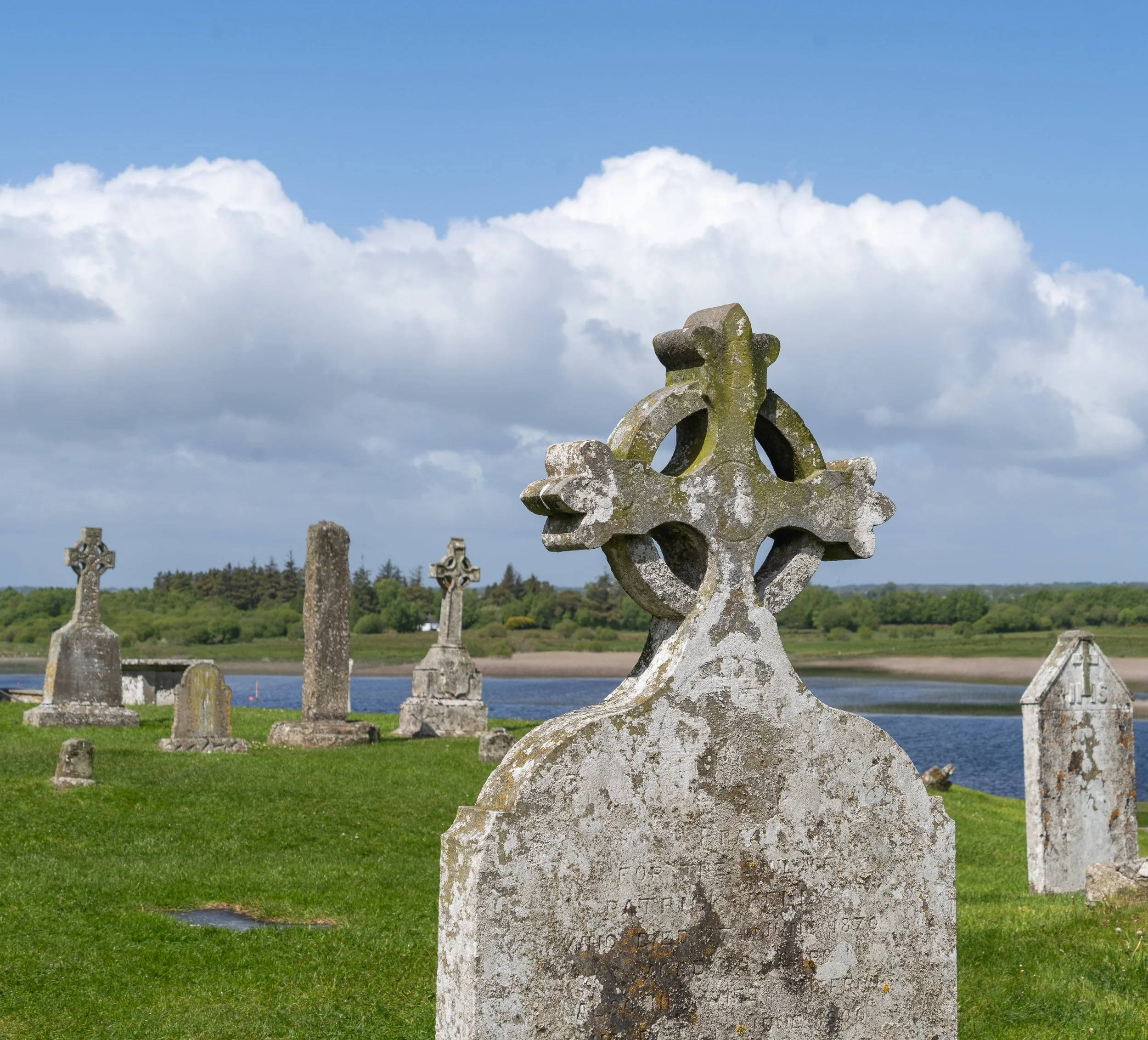 Historic Celtic cross standing in an Irish riverside cemetery beneath dramatic clouds, with ancient gravestones overlooking the water.
