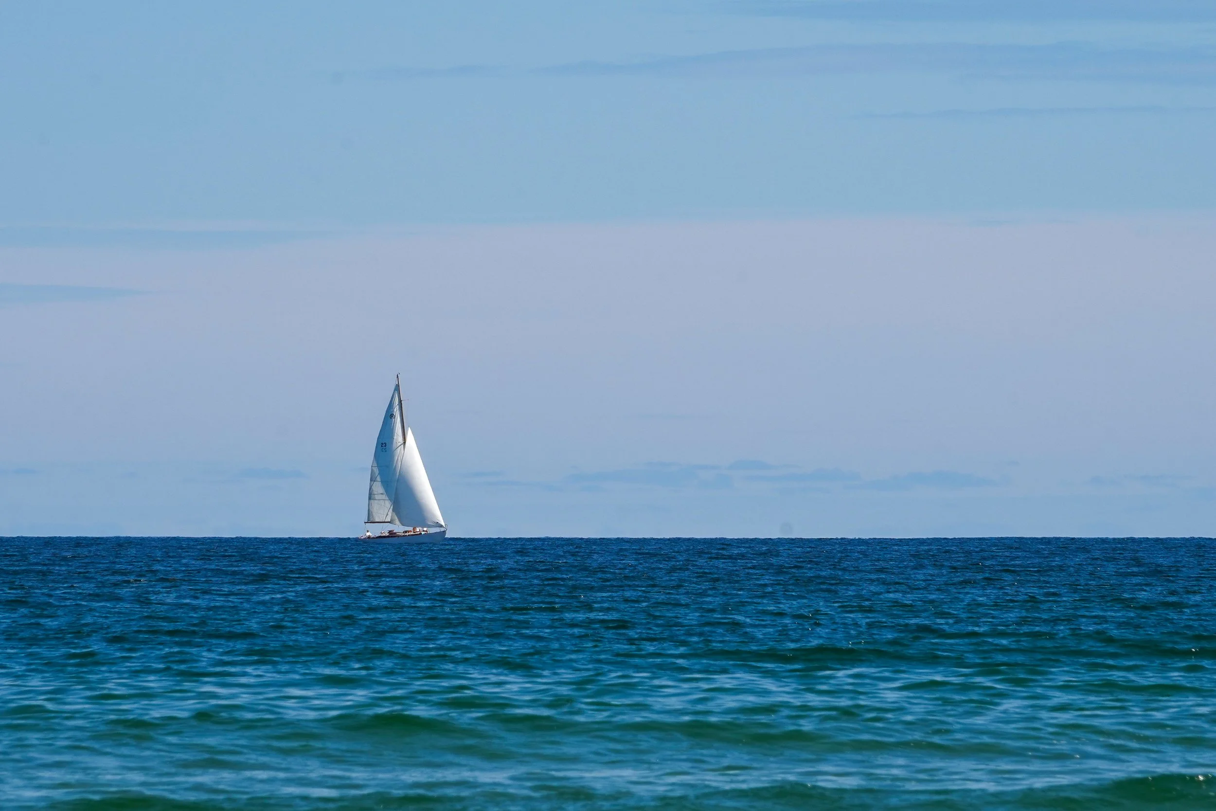 A solitary sailboat gliding across the Atlantic Ocean off the coast of Ogunquit, Maine, beneath a soft blue summer sky.