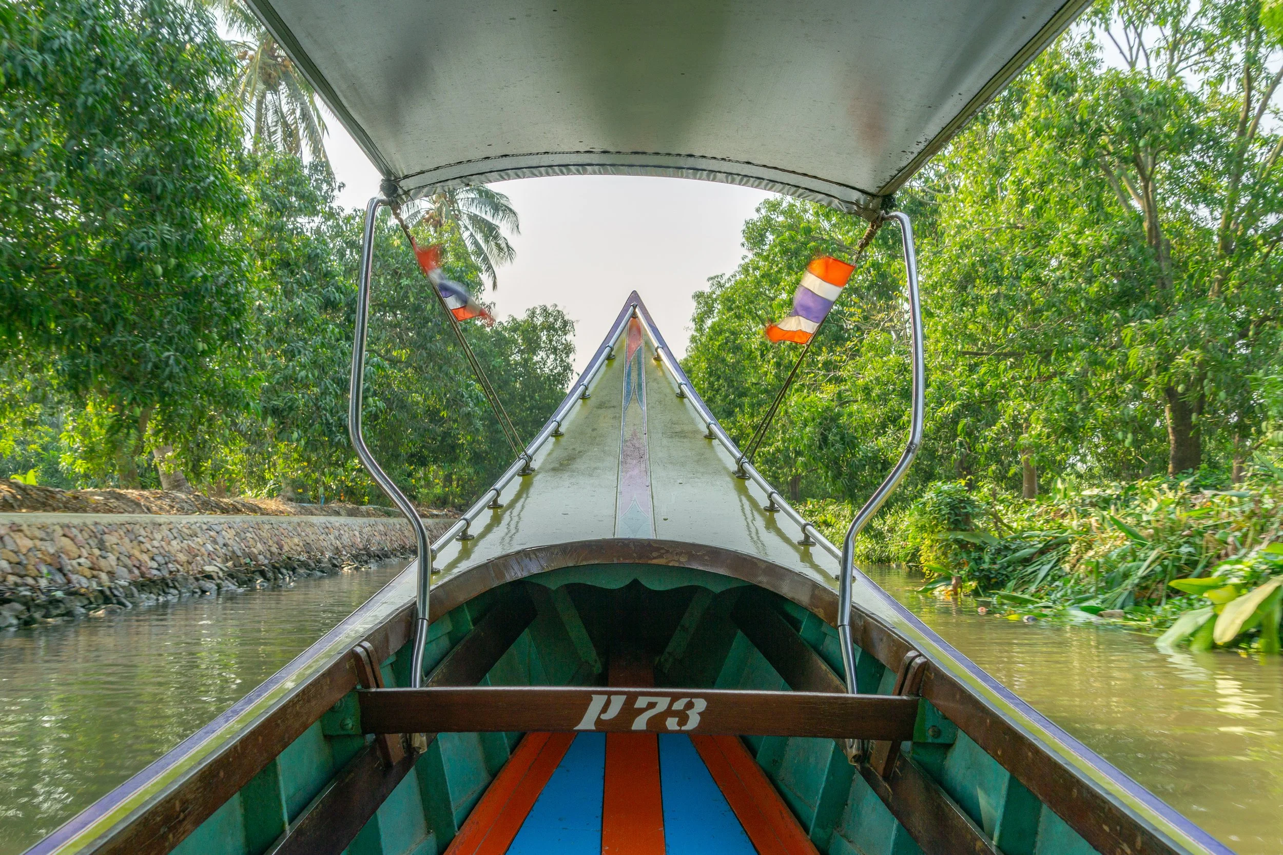 View from a traditional long-tail boat traveling along a tree-lined canal in Thailand with Thai flags at the bow.
