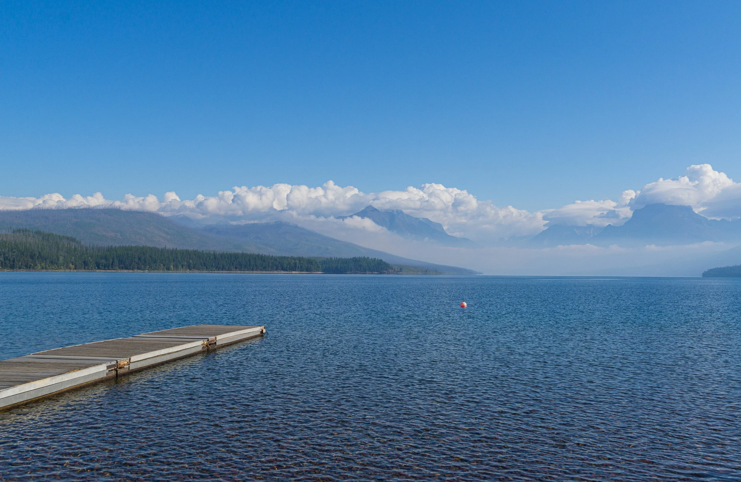 Calm blue lake with a small dock leading toward distant mountains and clouds in Glacier National Park.