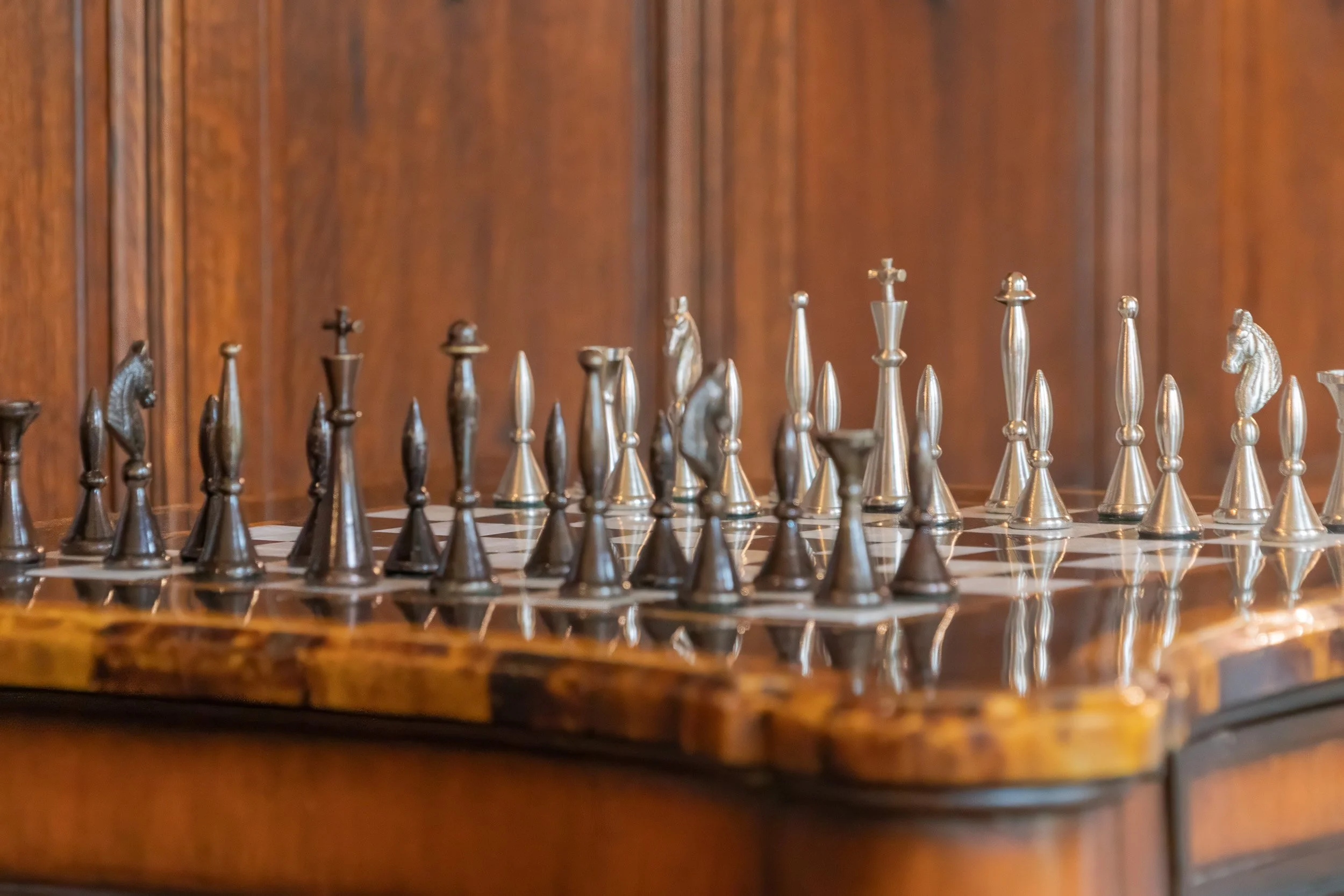 Metal chess pieces arranged on a polished chessboard against warm wooden paneling.