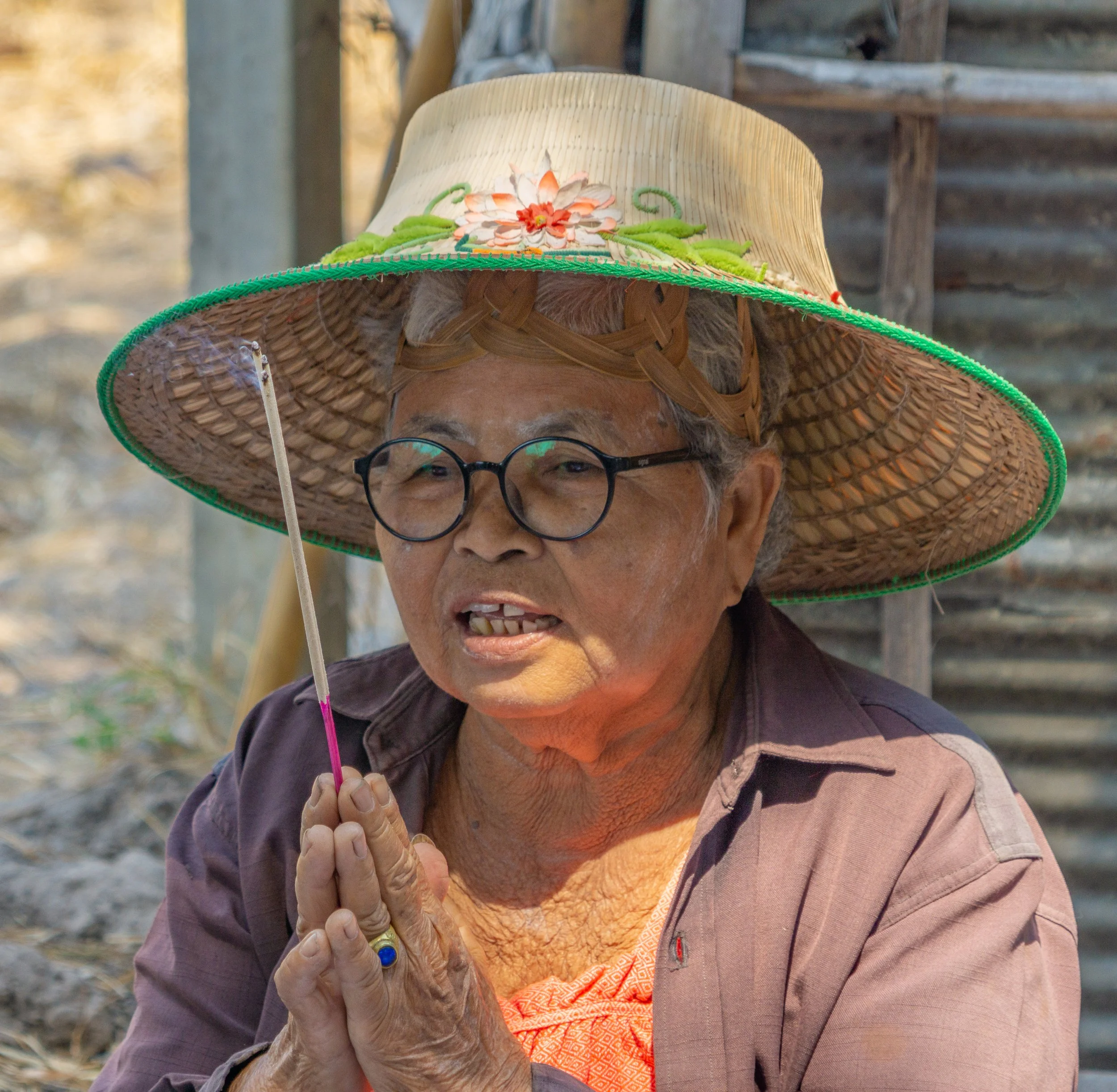Older woman holding a lit incense stick in prayer during a Buddhist ceremony in Thailand.