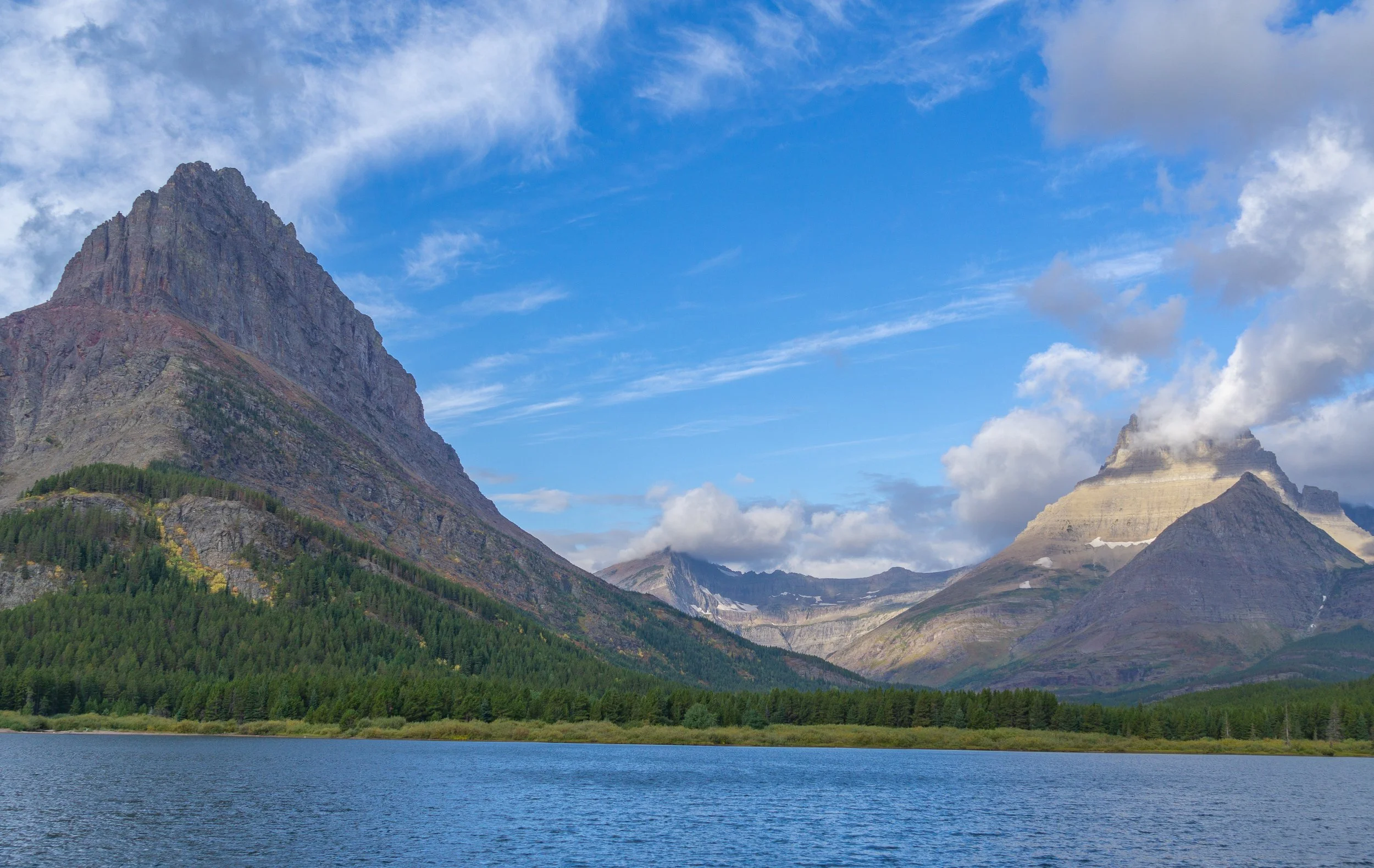Panoramic view of rugged mountains, evergreen forest, and a tranquil lake beneath dramatic clouds in Glacier National Park, Montana.